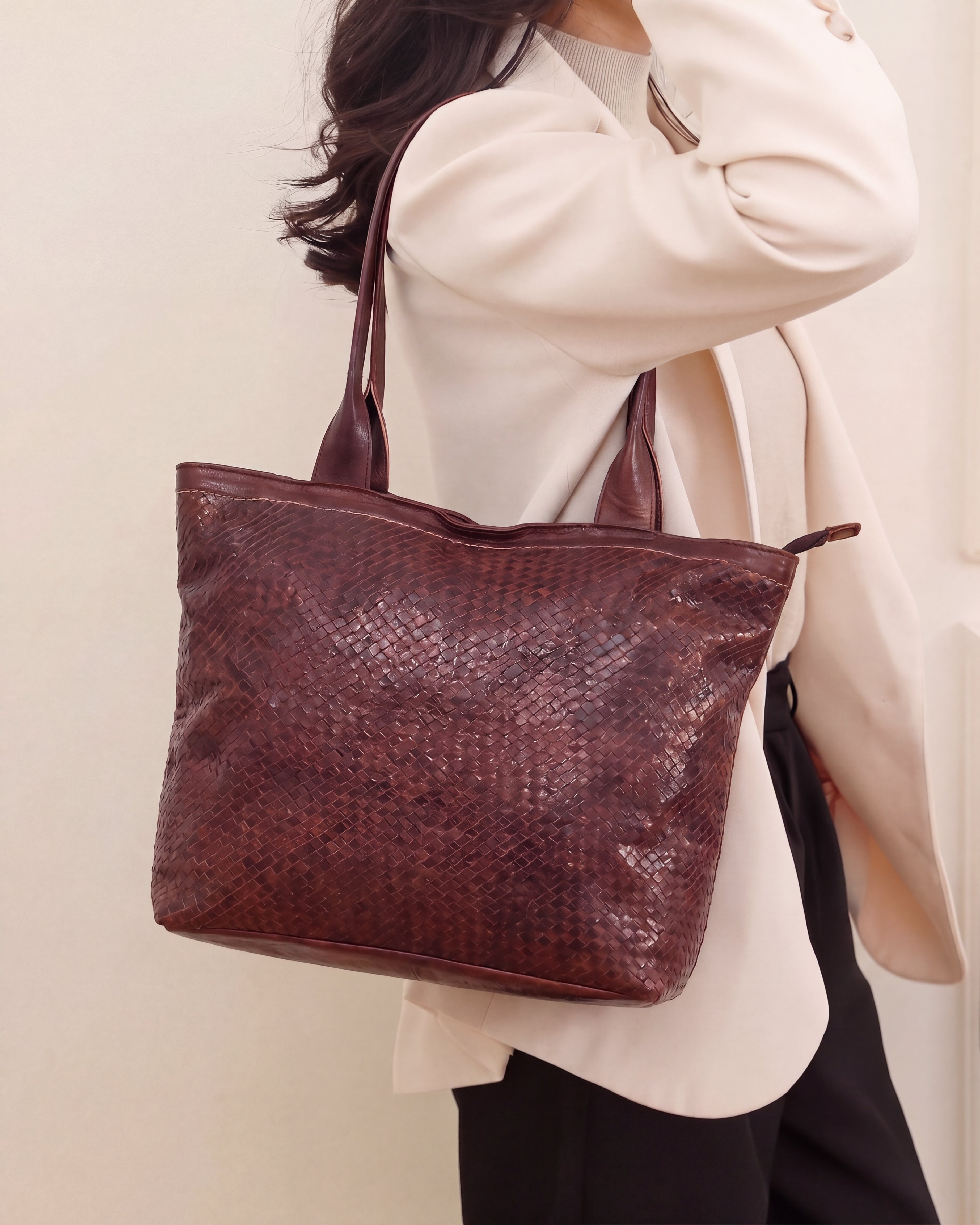 A person wearing a cream blazer and black pants carries a large, dark reddish-brown textured tote bag with shoulder straps.