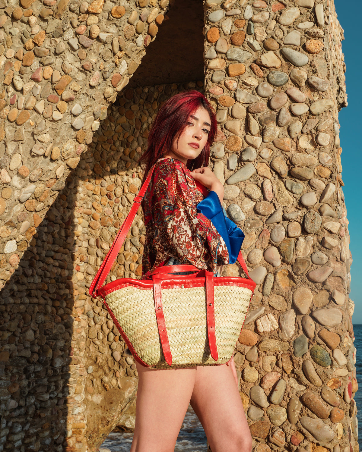 A woman with red hair stands in front of a stone archway, wearing a colorful patterned top and holding a large woven straw tote bag with red straps. The background features a textured stone wall and a sliver of blue sky.