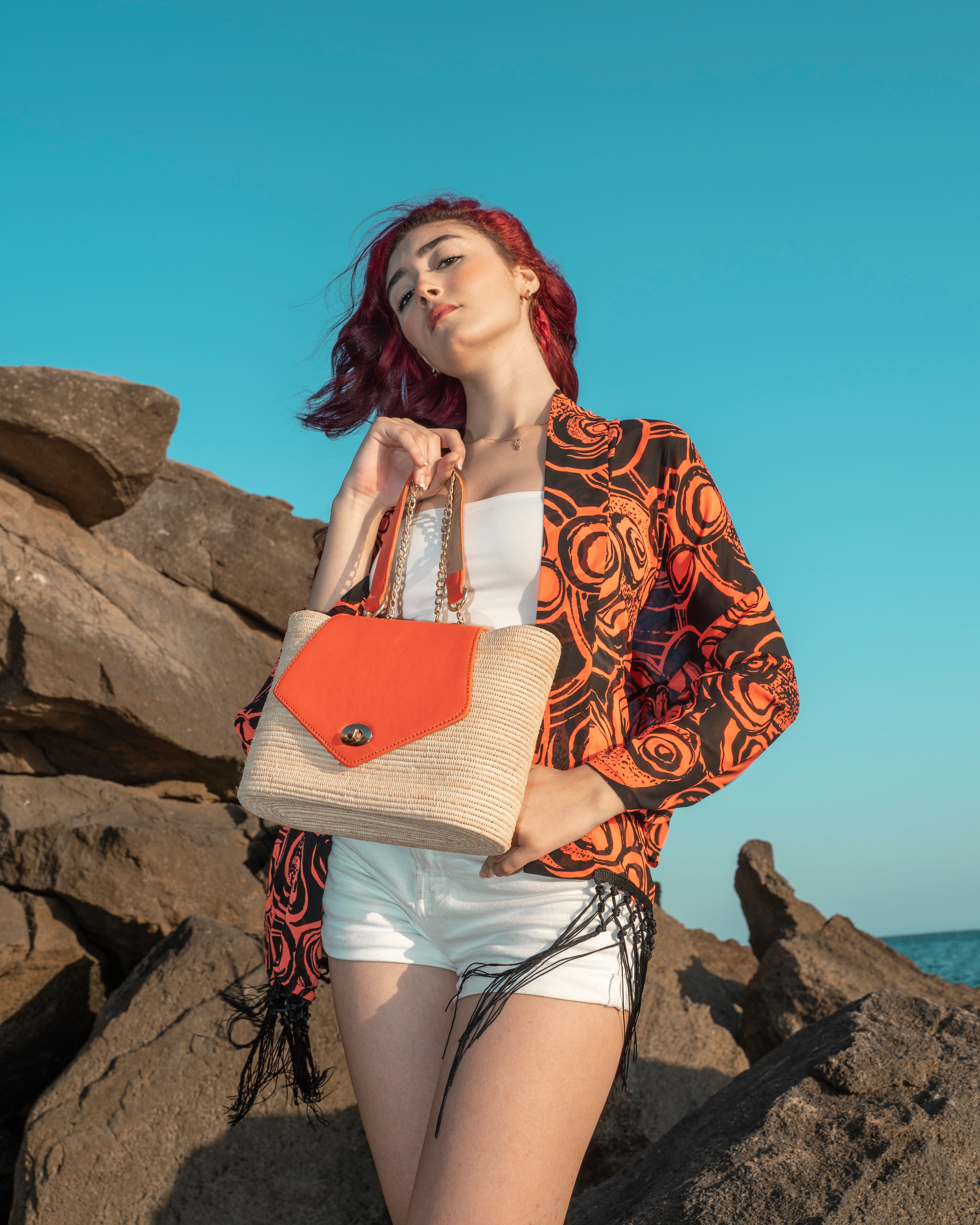 A woman with red hair stands on rocky outdoor ground holding a woven straw tote bag with a bright orange front flap and orange handles. She wears a white strapless top, white shorts, and an orange-and-black patterned cardigan against a blue sky.