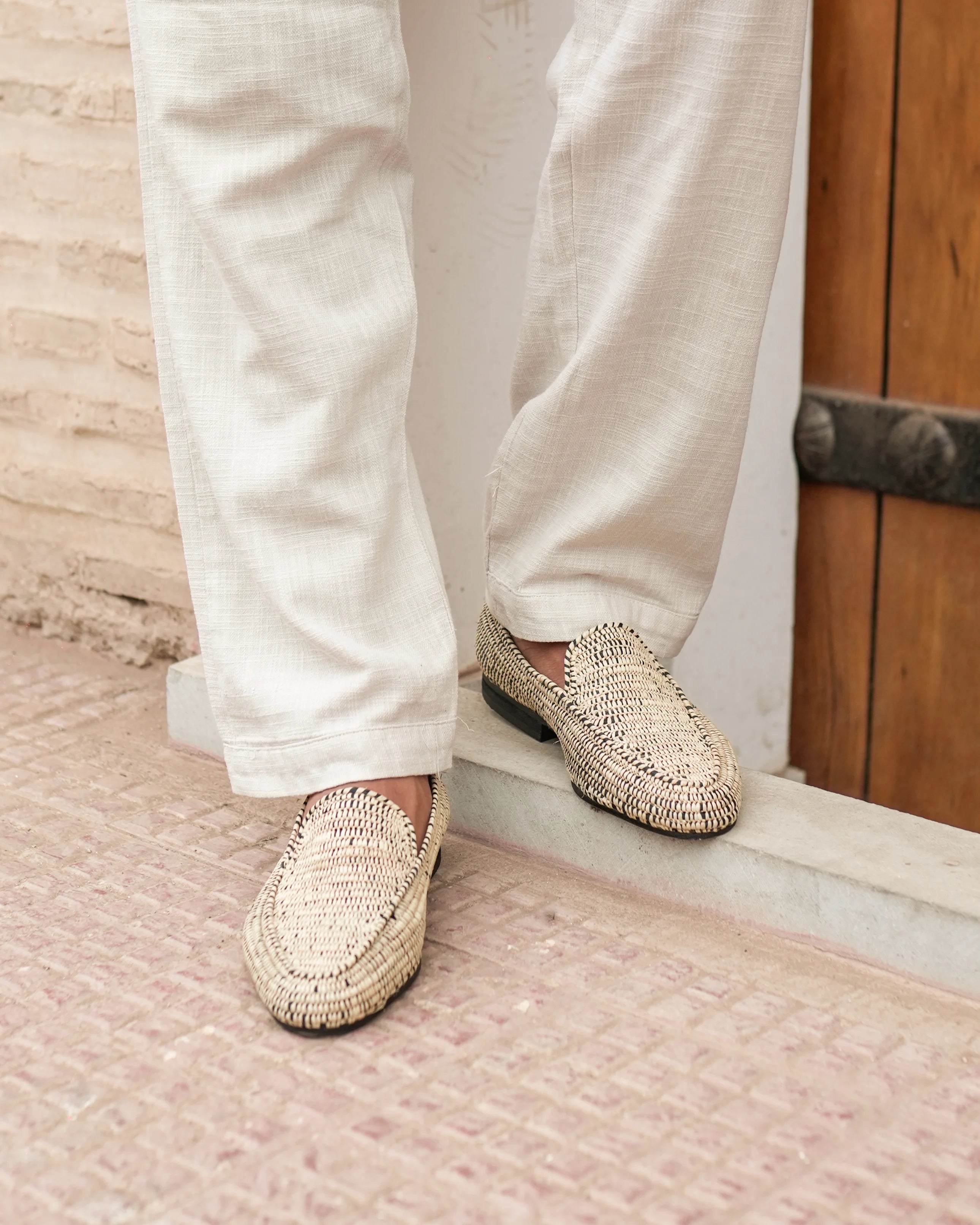 Person wearing beige woven loafers with white pants standing on a textured floor.