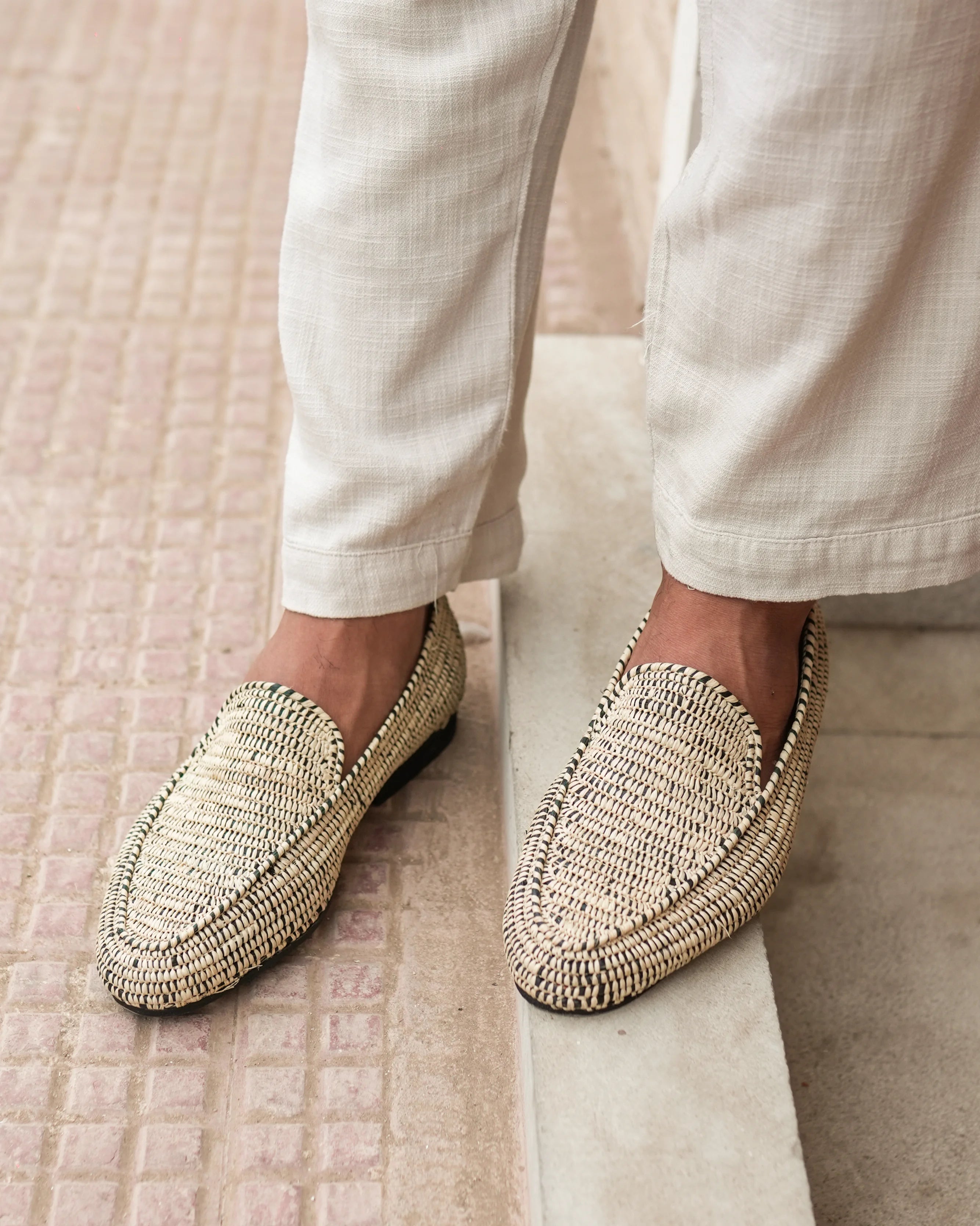 Beige woven loafers worn with white pants on a textured floor.