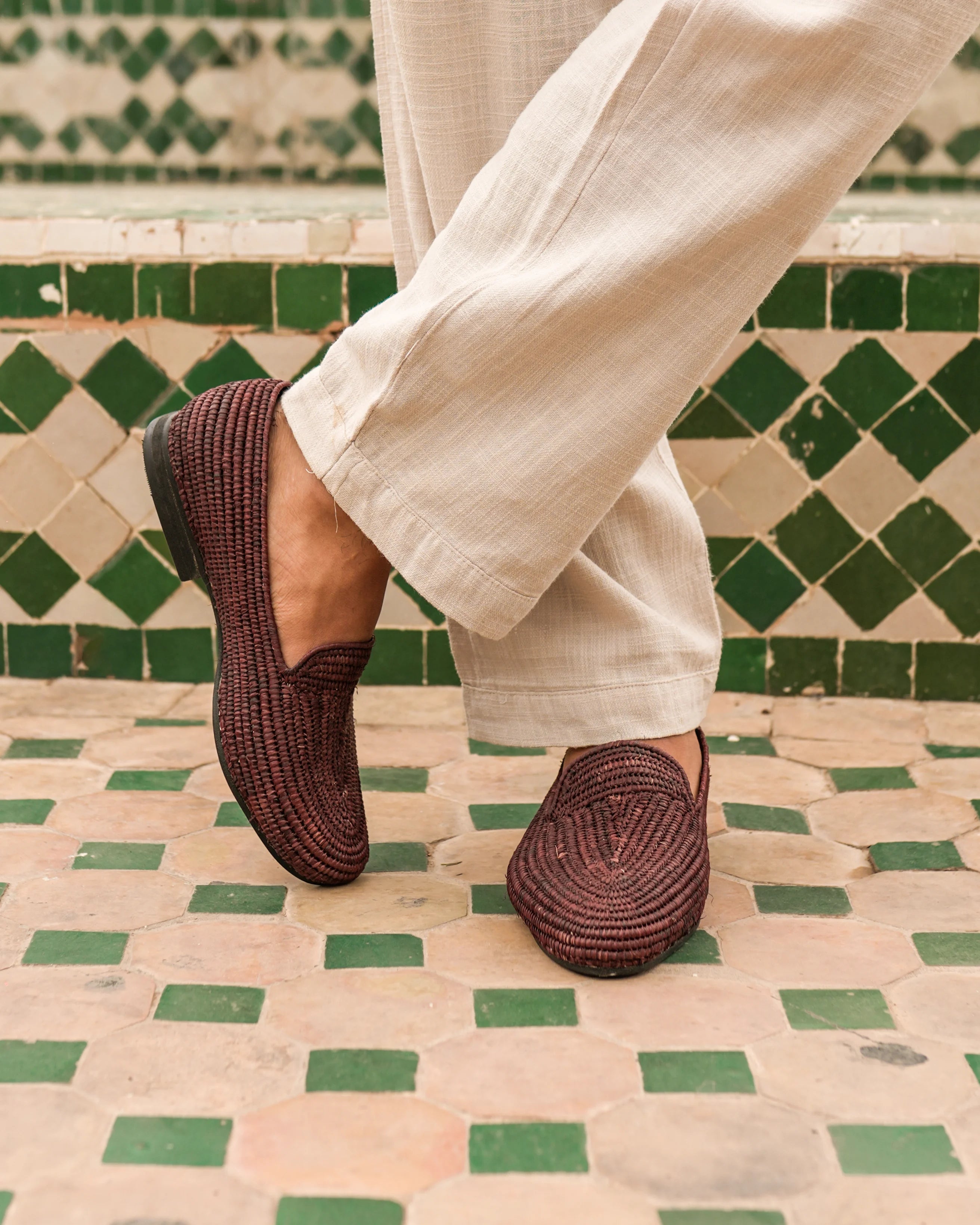 Brown woven shoes worn with light-colored pants on a green and beige tiled floor.