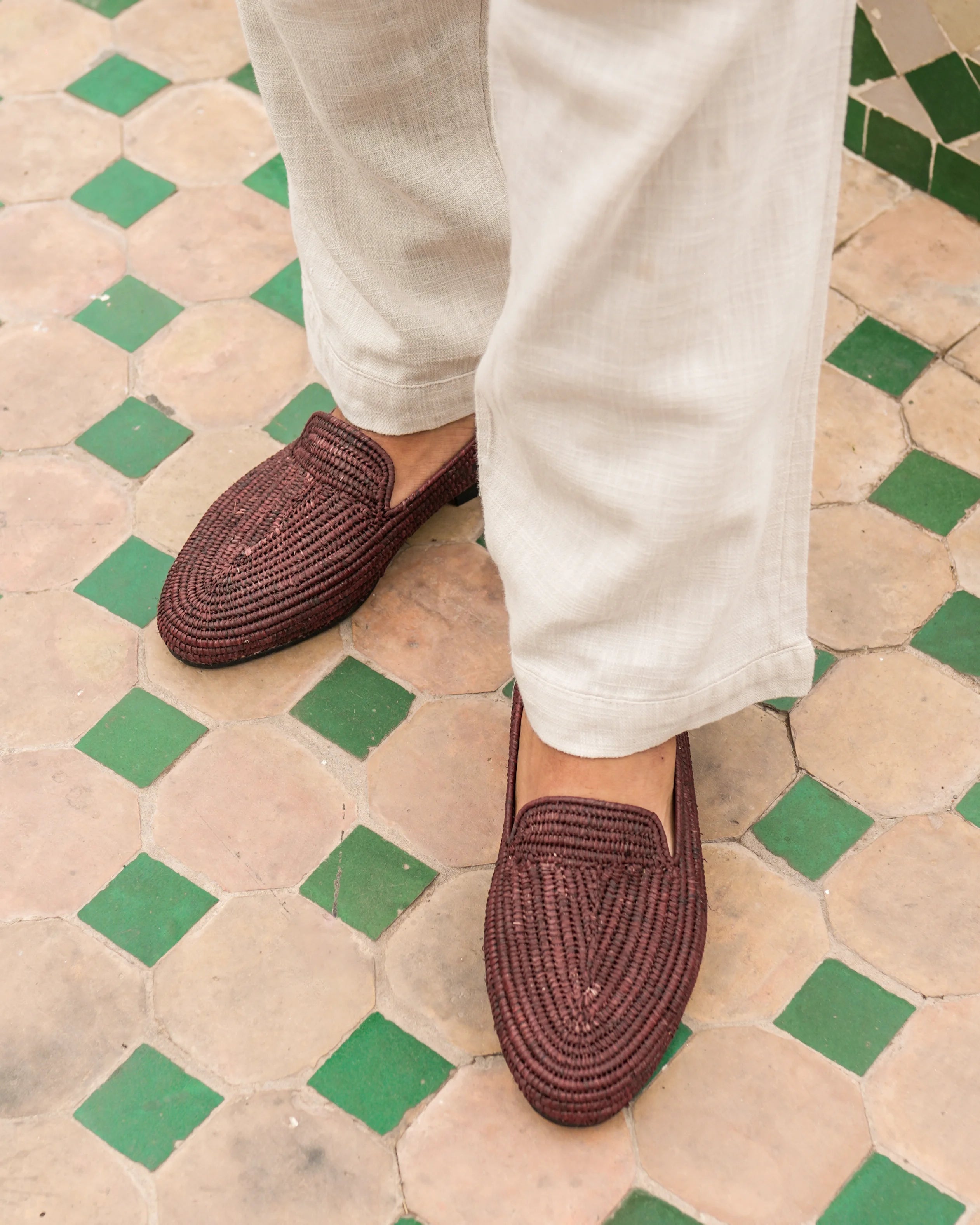 Brown woven loafers worn with white pants on a patterned floor.