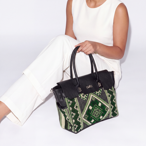 A woman in a white sleeveless top and white pants holds a structured handbag with black leather trim and handles, featuring a green and black geometric patterned body, against a clean white background.