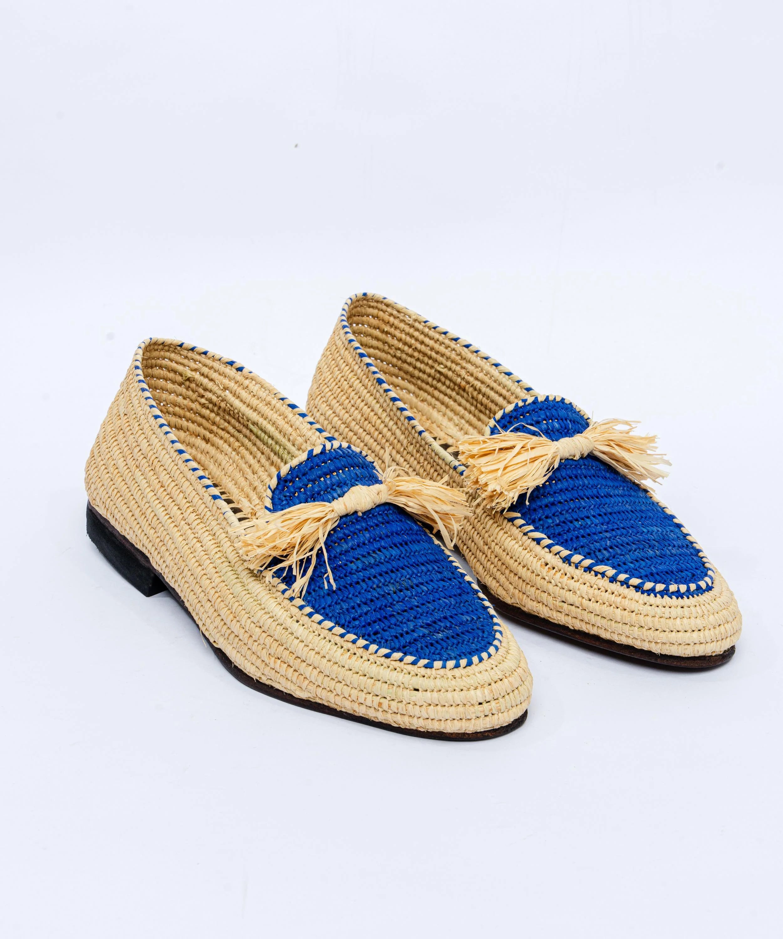 Pair of raffia loafers with blue insoles and tassels on a white background