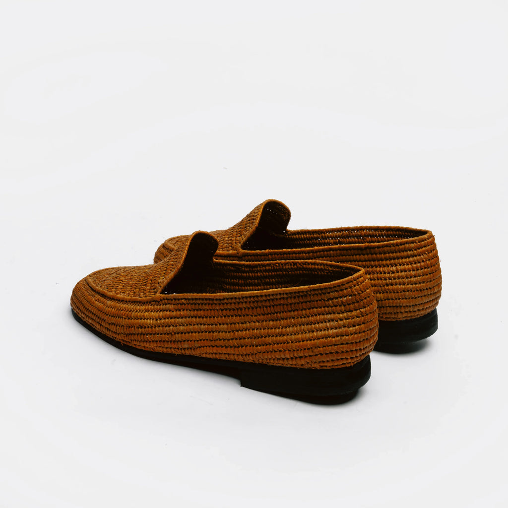 A pair of brown woven straw loafers with black soles, shown from a side-rear angle on a clean white background.