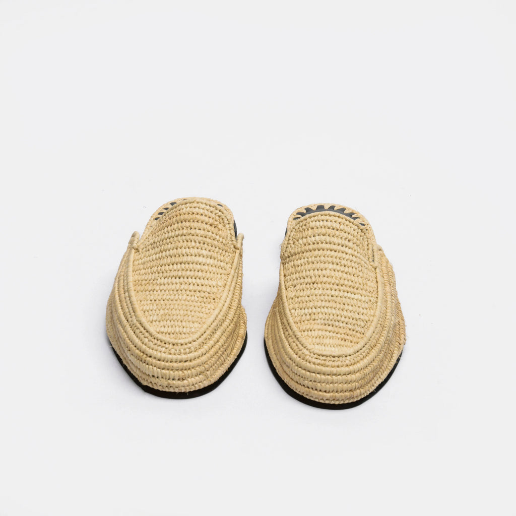 A pair of natural straw woven slip-on loafers with black soles, photographed from a top-down angle on a light gray background.