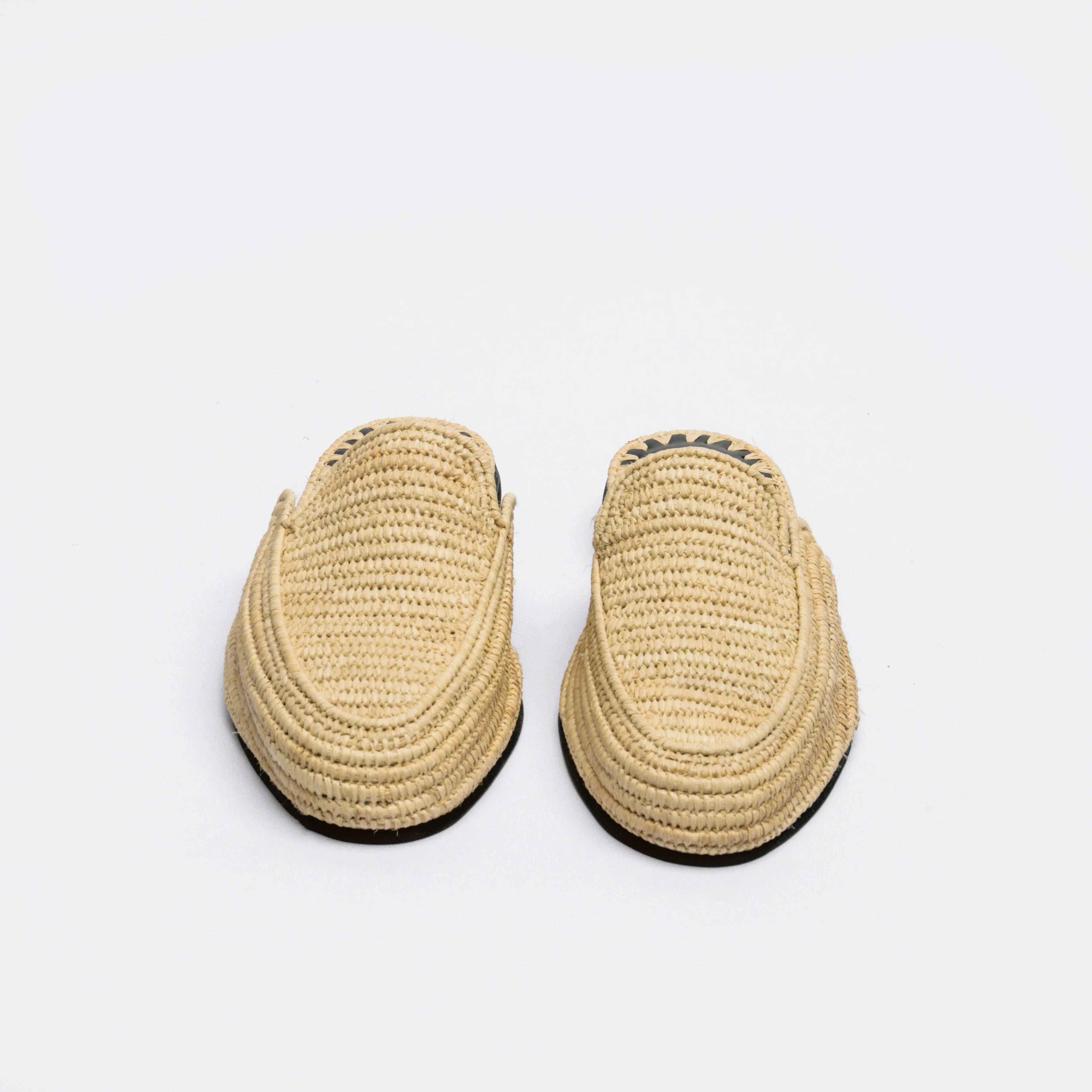 A pair of natural straw woven slip-on loafers with black soles, photographed from a top-down angle on a light gray background.