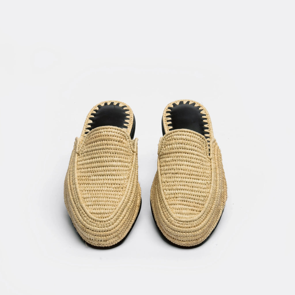A pair of woven raffia slip-on mule sandals with black soles and black interior lining, shown from a top-down view on a light gray background.