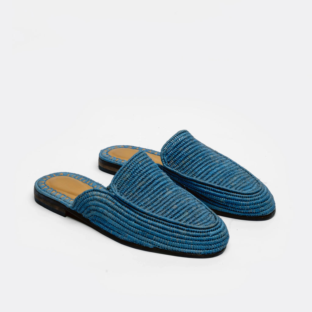A pair of blue woven raffia mule slippers with black soles and tan insoles, displayed on a clean white background.