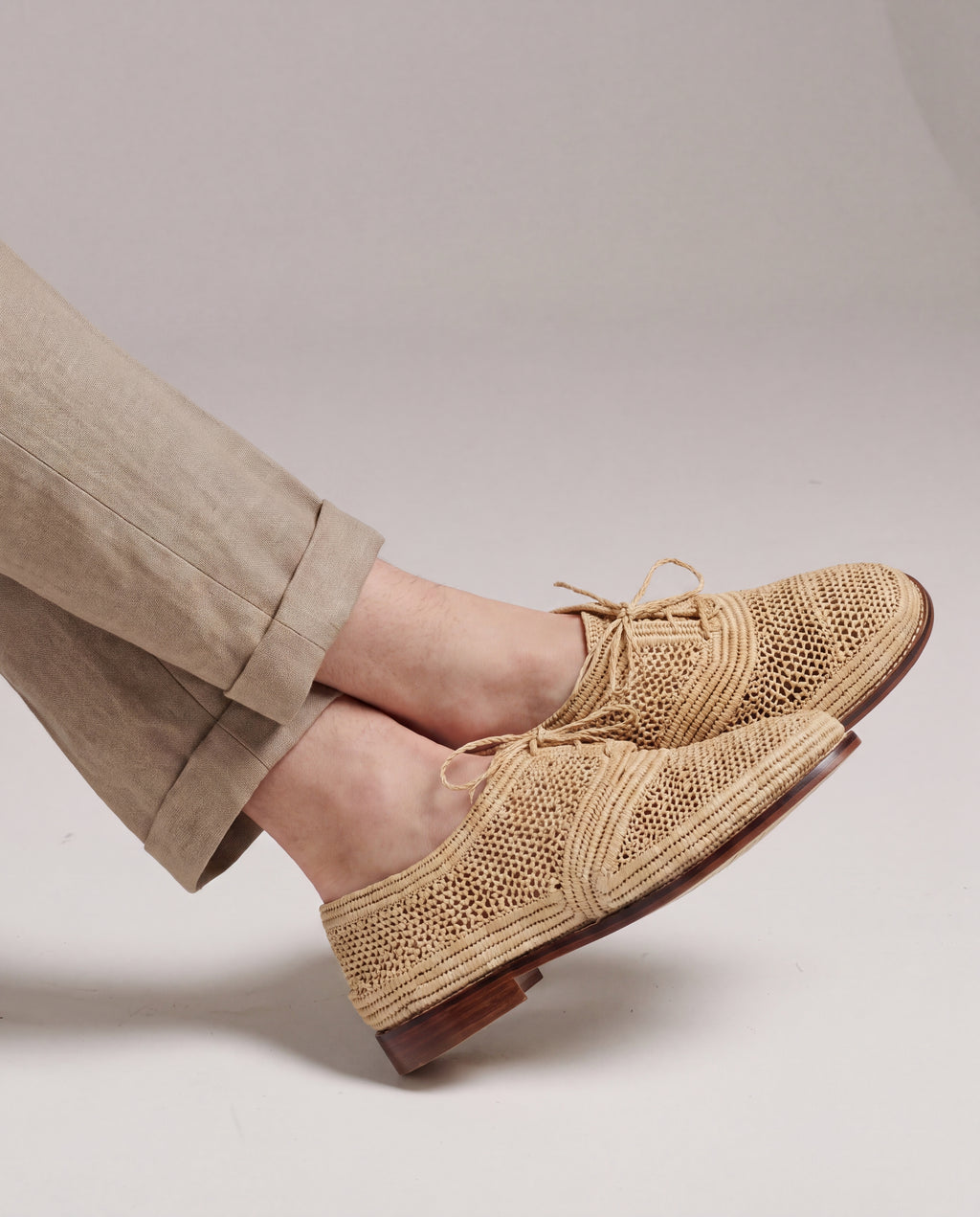 A men wearing beige woven lace-up shoes with tan trousers, shown from the calves down against a plain light gray studio background.