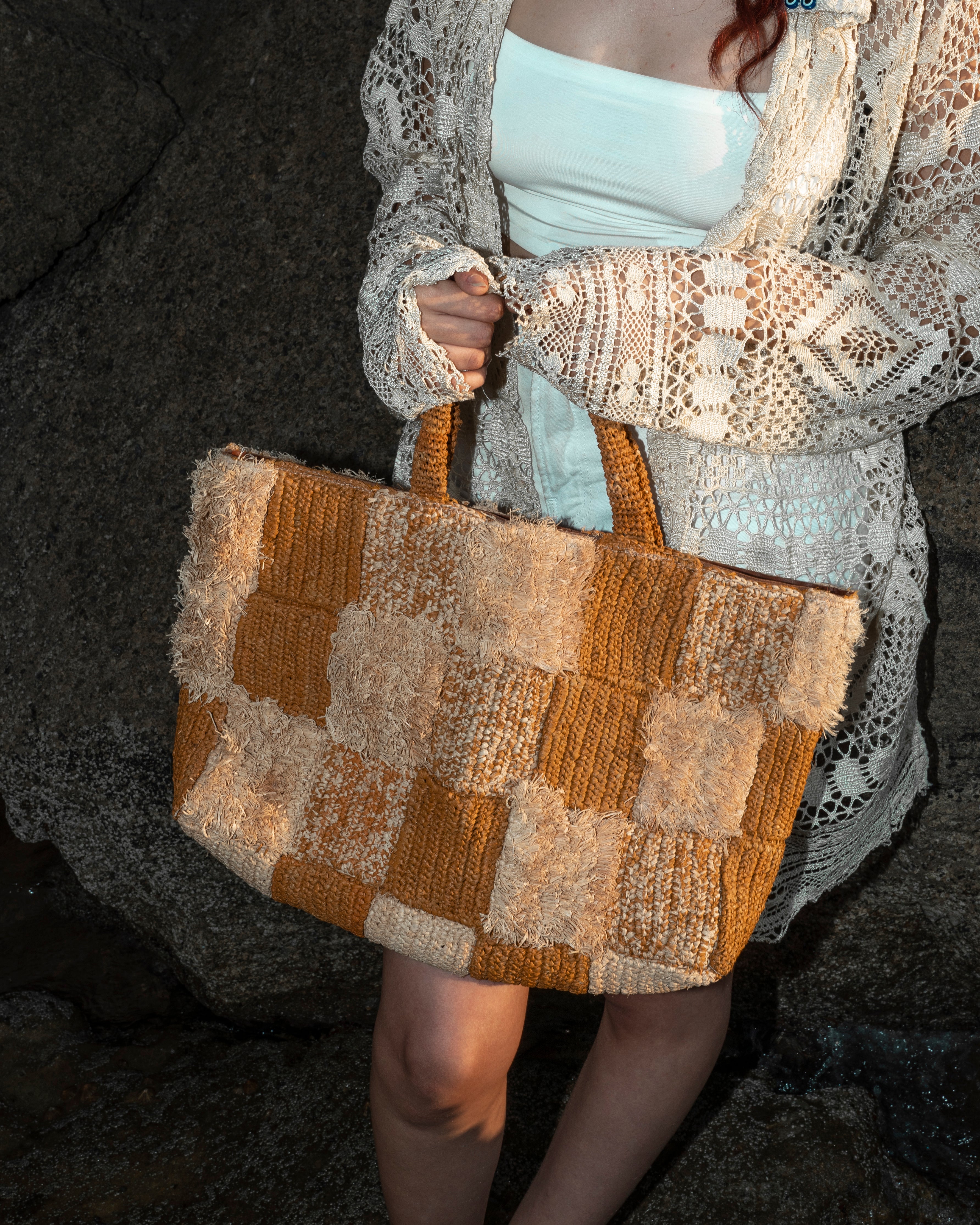 A person wearing a cream crochet cardigan and white tube top holds a large patchwork knit tote bag in mustard orange and cream against a dark rocky background.