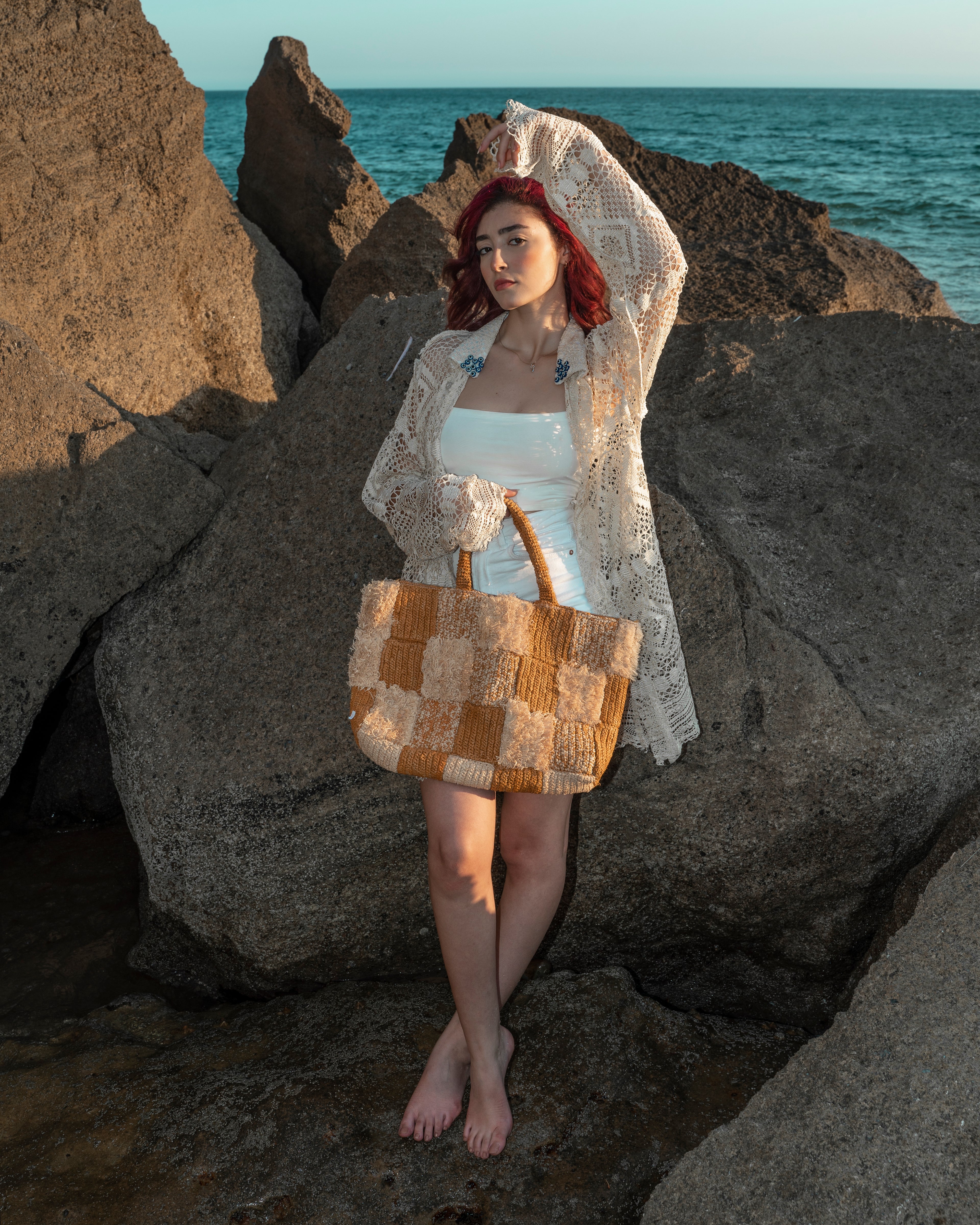 A woman stands barefoot on large rocks by the ocean, wearing a white bikini, a cream crochet cardigan, and holding a woven straw tote bag with tan and cream checkered panels.