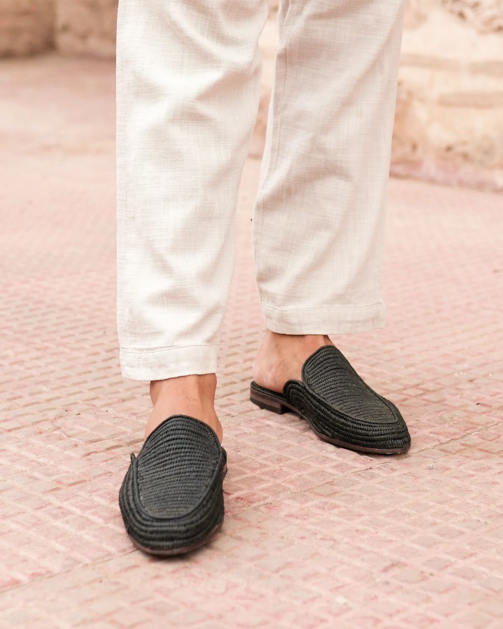 Person wearing black slip-on shoes with white pants on a textured pink surface