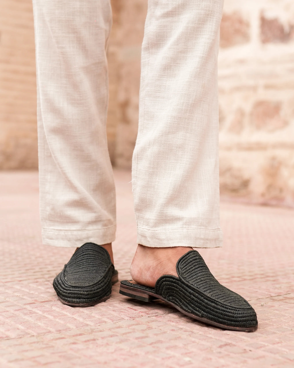 Black slip-on shoes worn with light-colored pants on a textured surface.