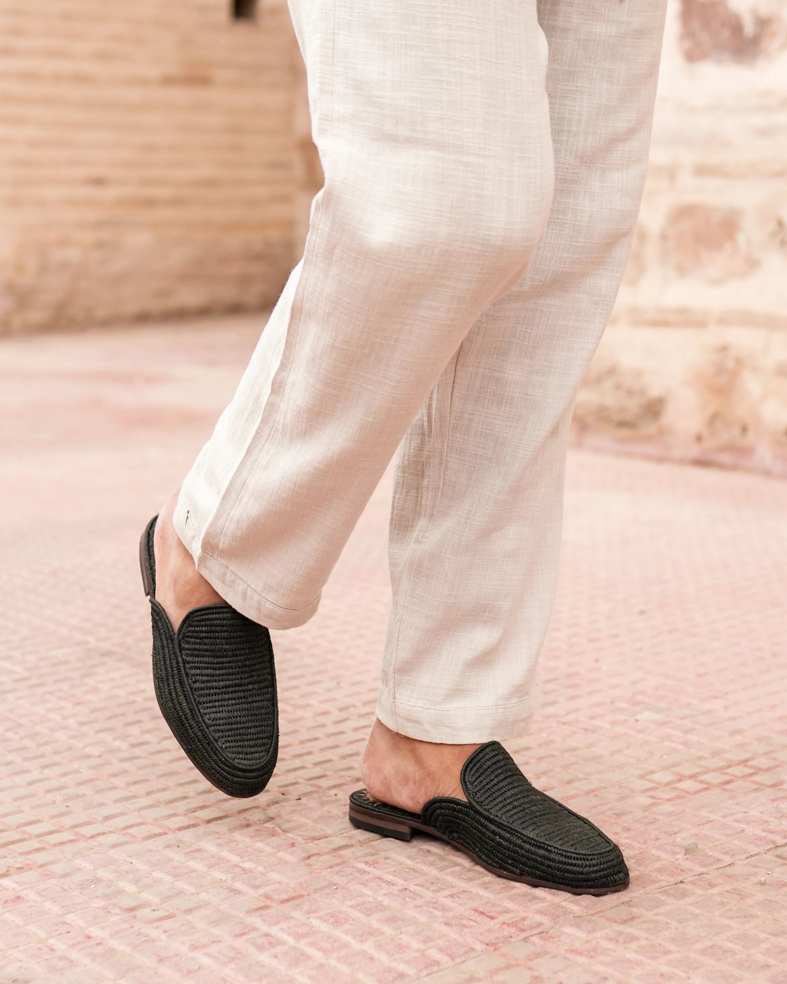 Person wearing black slip-on shoes with white pants on a textured pink surface