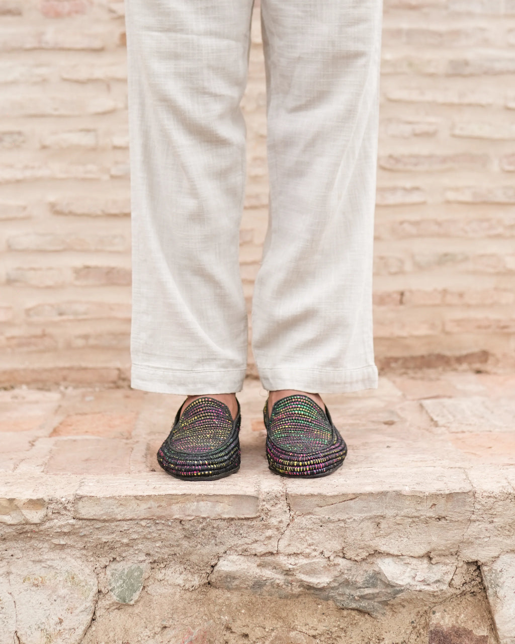 Person wearing white pants and colorful sandals standing on a stone surface with a wooden wall background.