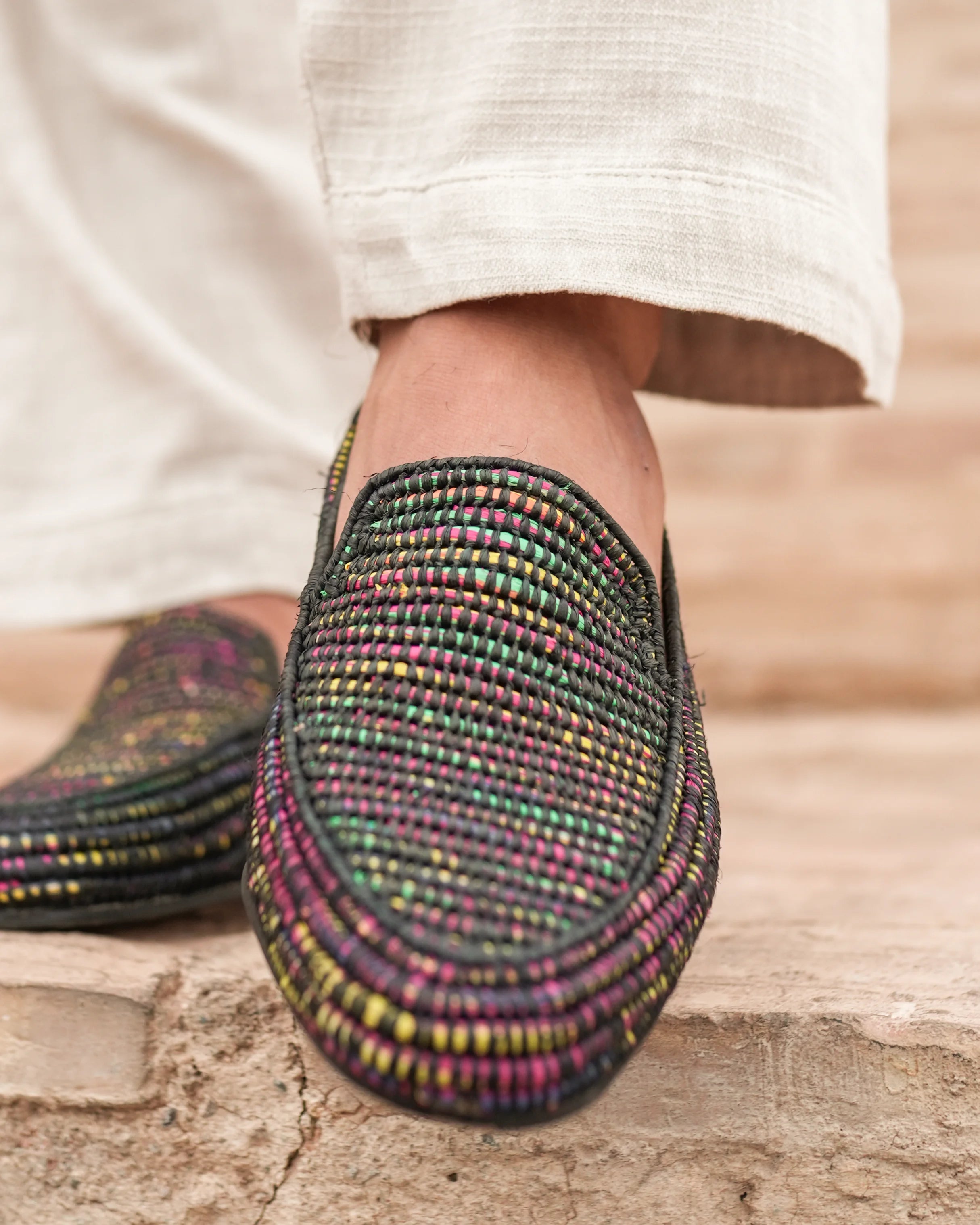 Multicolored woven shoes worn with light-colored pants on a wooden floor.