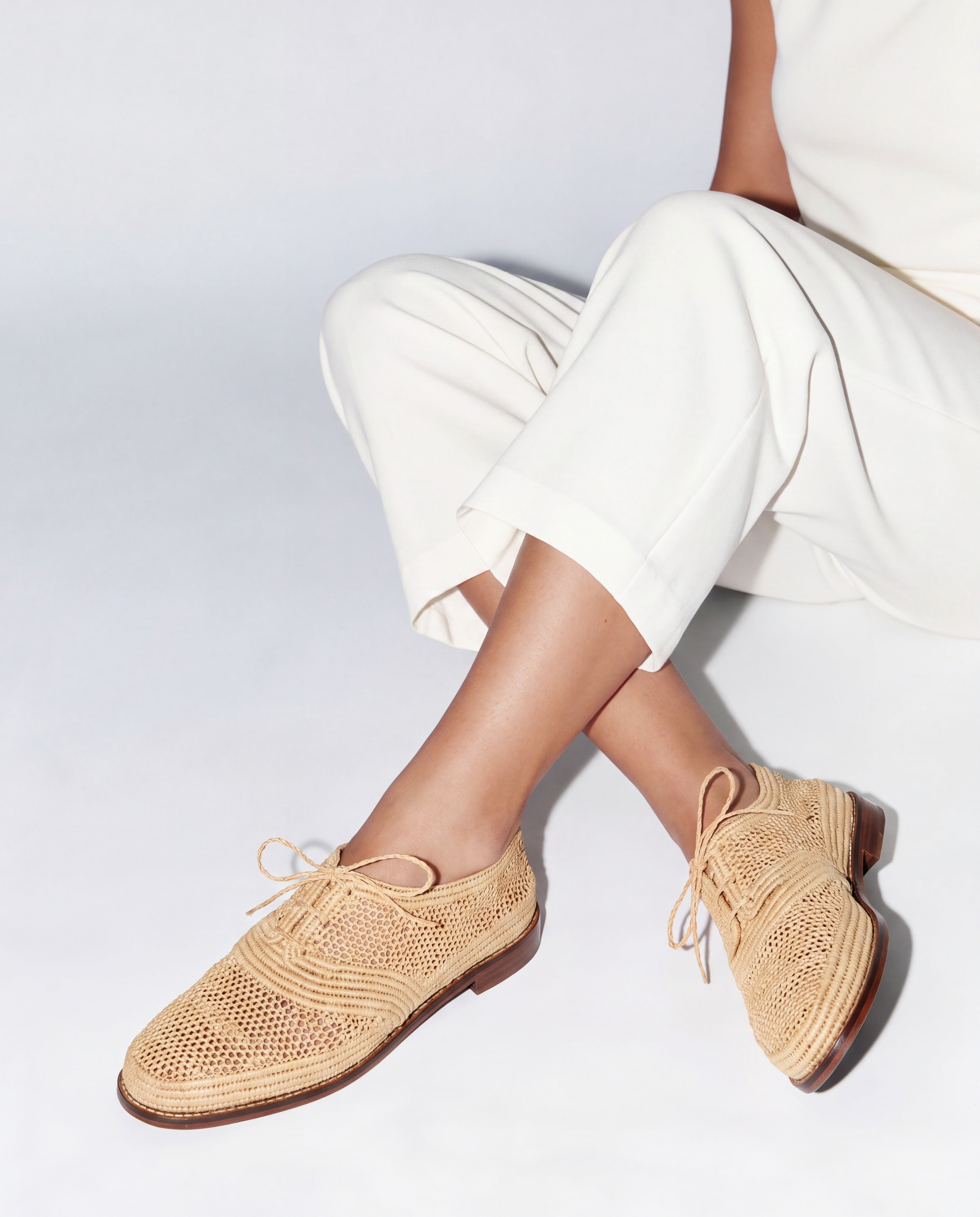 A person seated against a light gray studio background wearing woven beige lace-up shoes and off-white cropped pants.