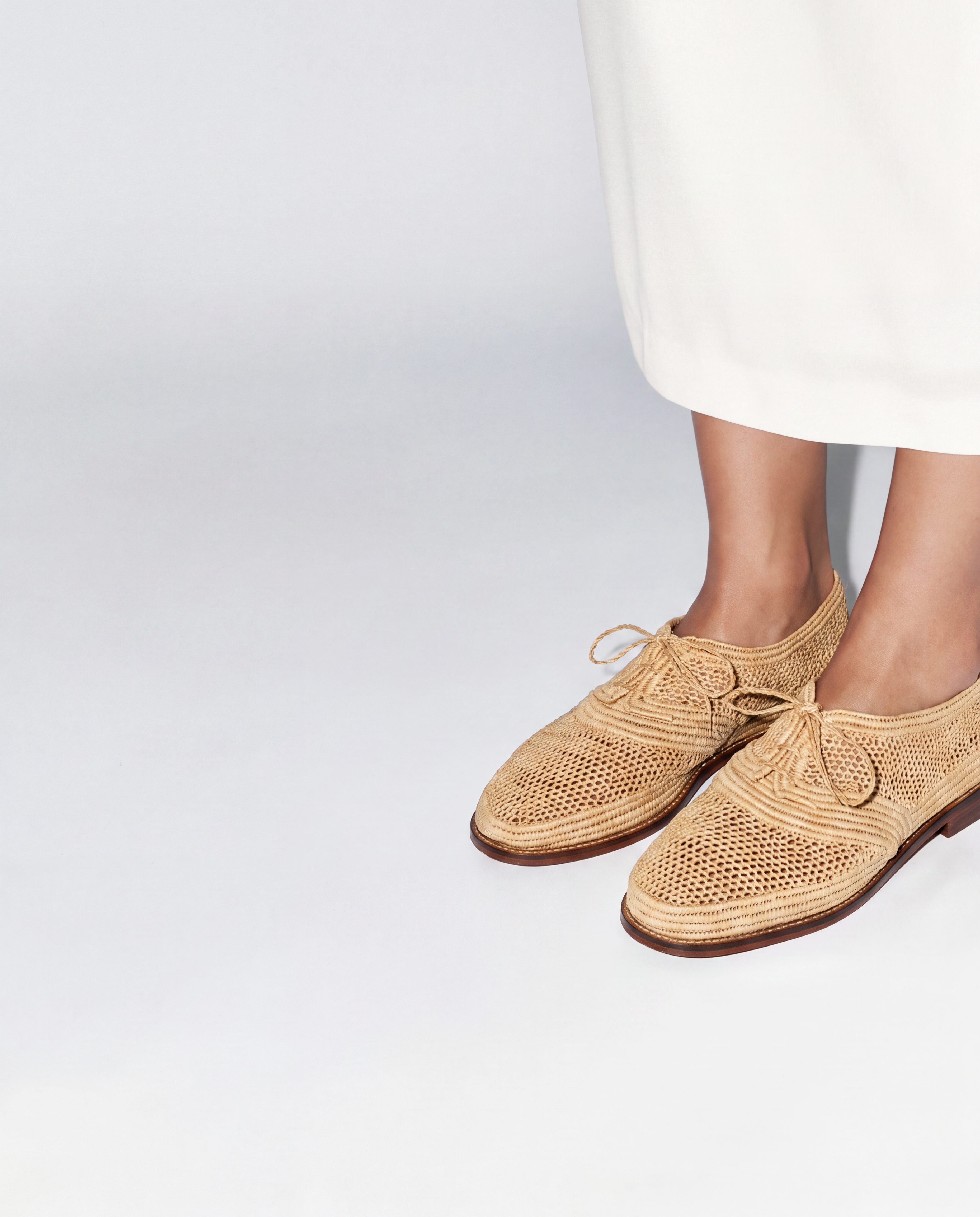 A person wearing beige woven lace-up oxford shoes with a white skirt, shown from the knees down against a clean white studio background.