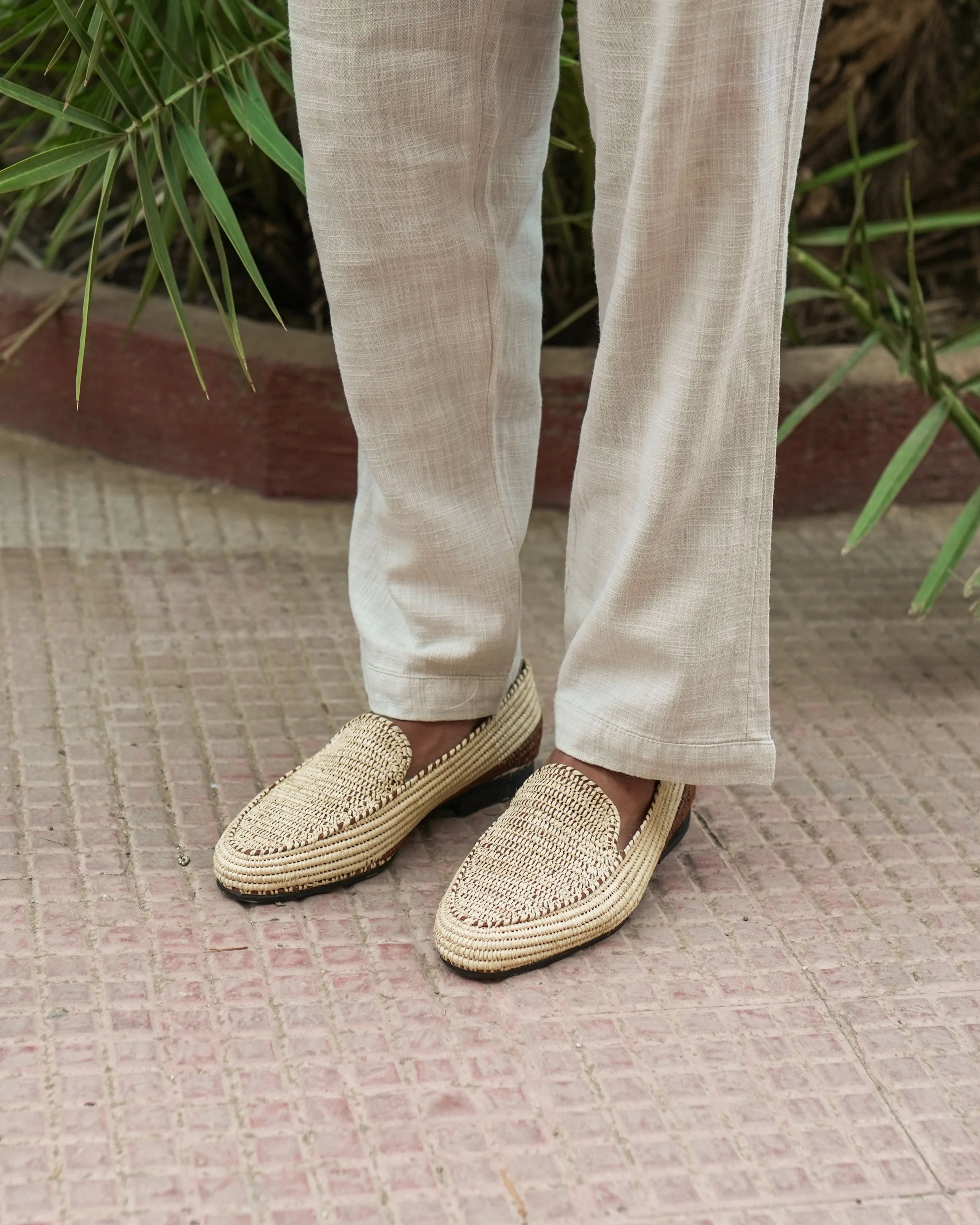 Person wearing beige woven shoes and light-colored pants on a textured surface with greenery in the background.