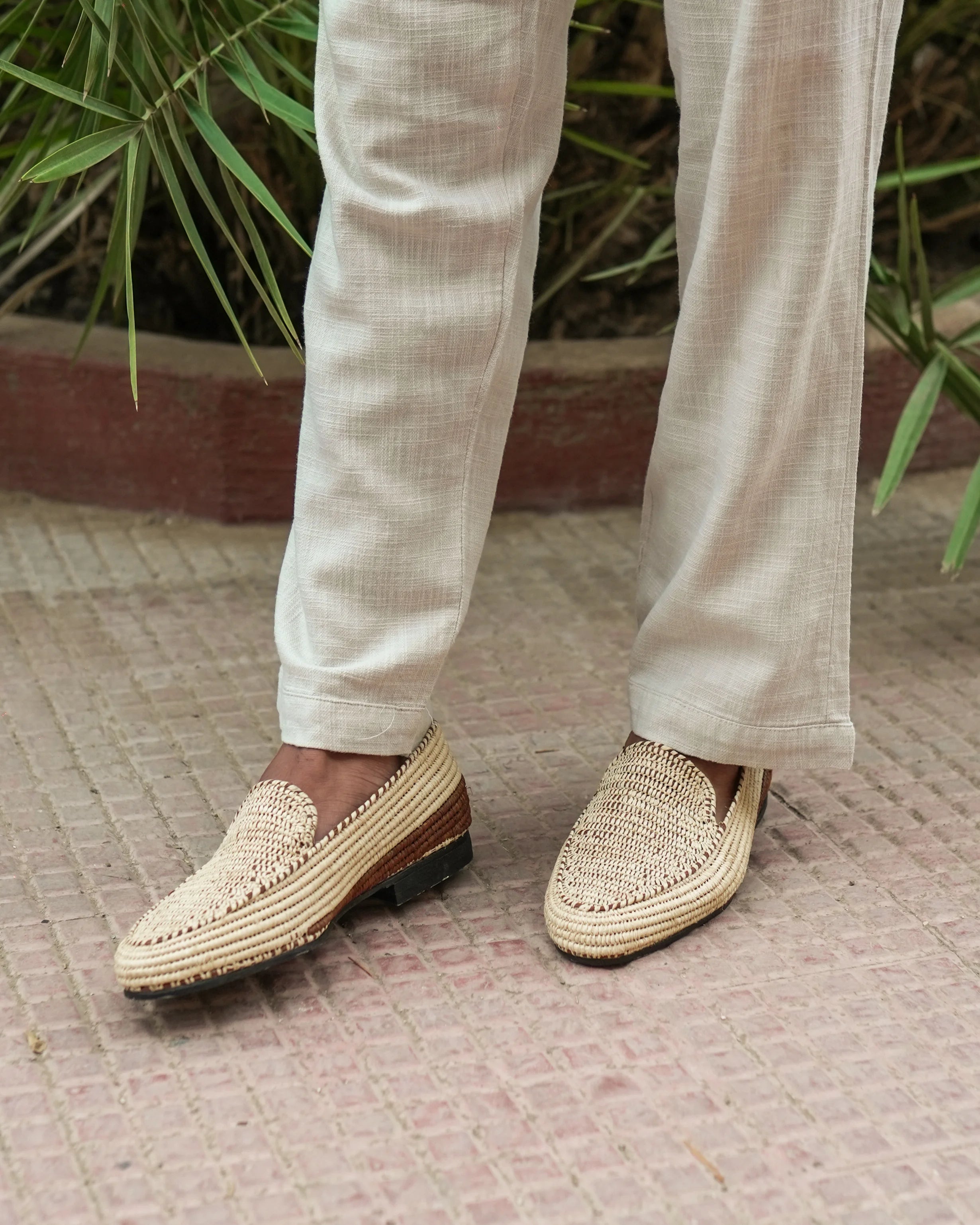 Person wearing white pants and woven shoes on a patterned floor with plants in the background