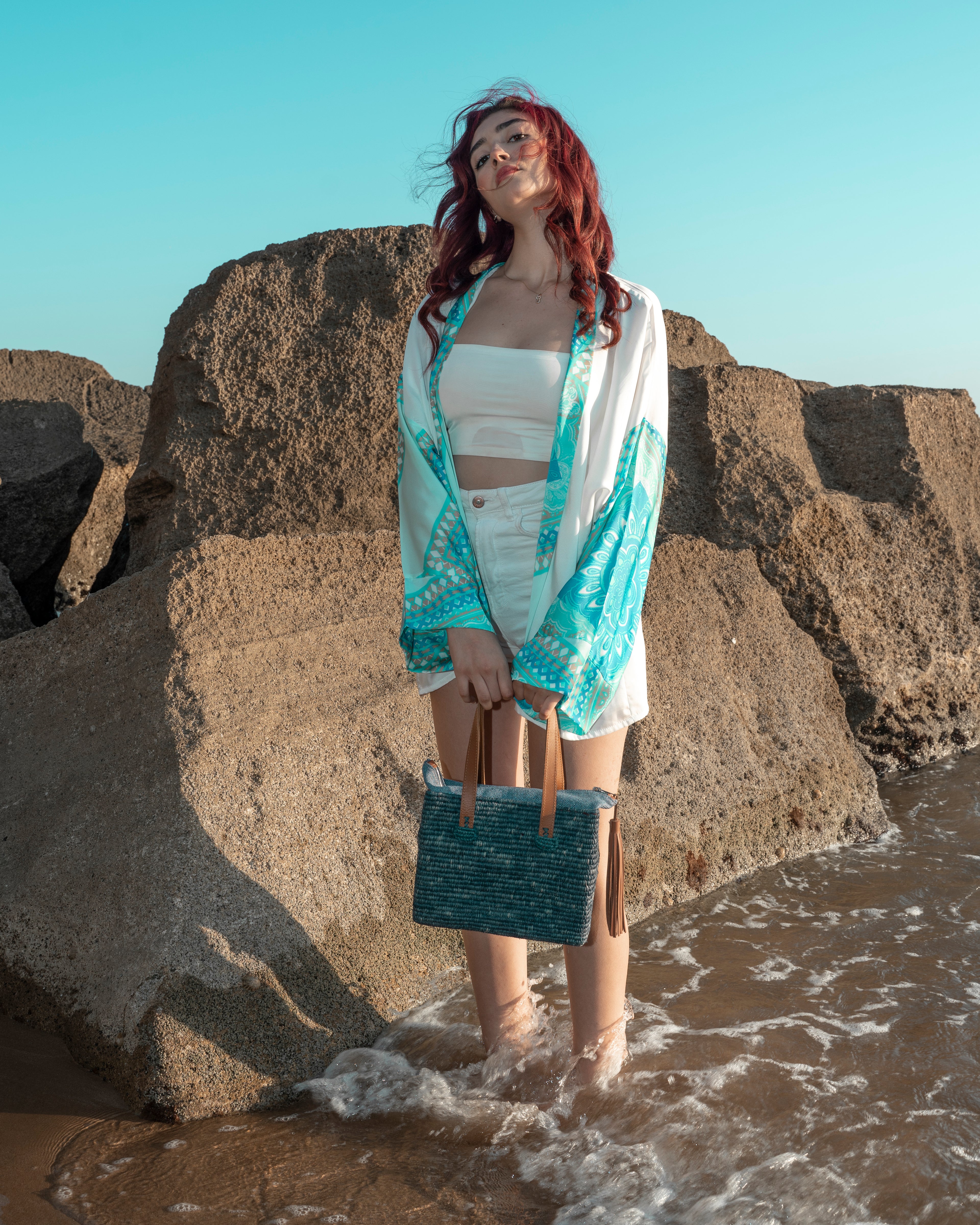 A woman with red hair stands in shallow water among large rocks, wearing a white crop top, white shorts, a white open-front cover-up with turquoise and white patterned sleeves, and holding a turquoise woven tote bag.