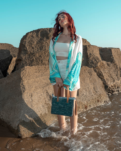 A woman with red hair stands in shallow water among large rocks, wearing a white crop top, white shorts, a white open-front cover-up with turquoise and white patterned sleeves, and holding a turquoise woven tote bag.