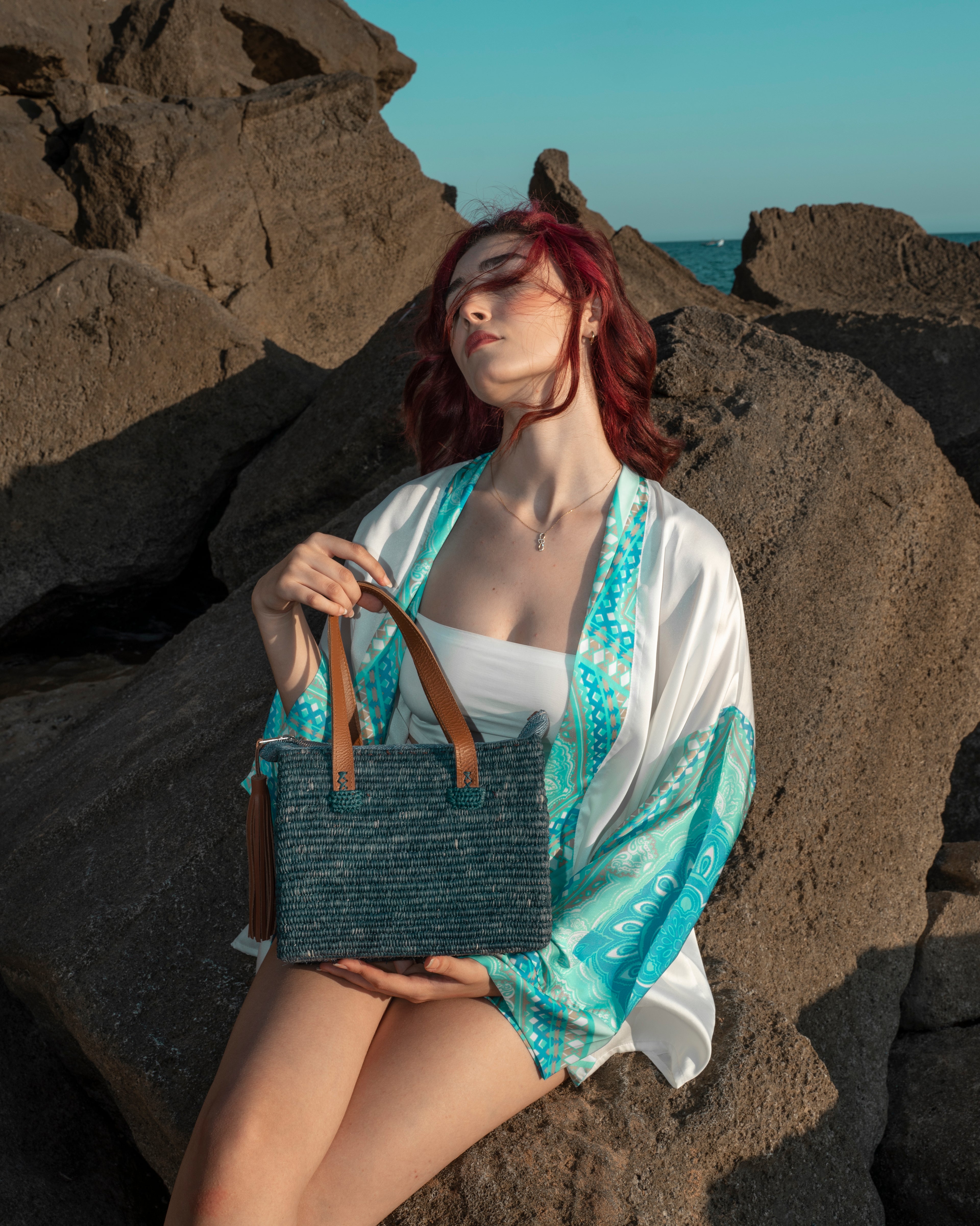 A woman with red hair sits on large rocks outdoors near the ocean, wearing a white and turquoise patterned kimono-style cover-up over a white top. She holds a dark woven tote bag with brown handles.