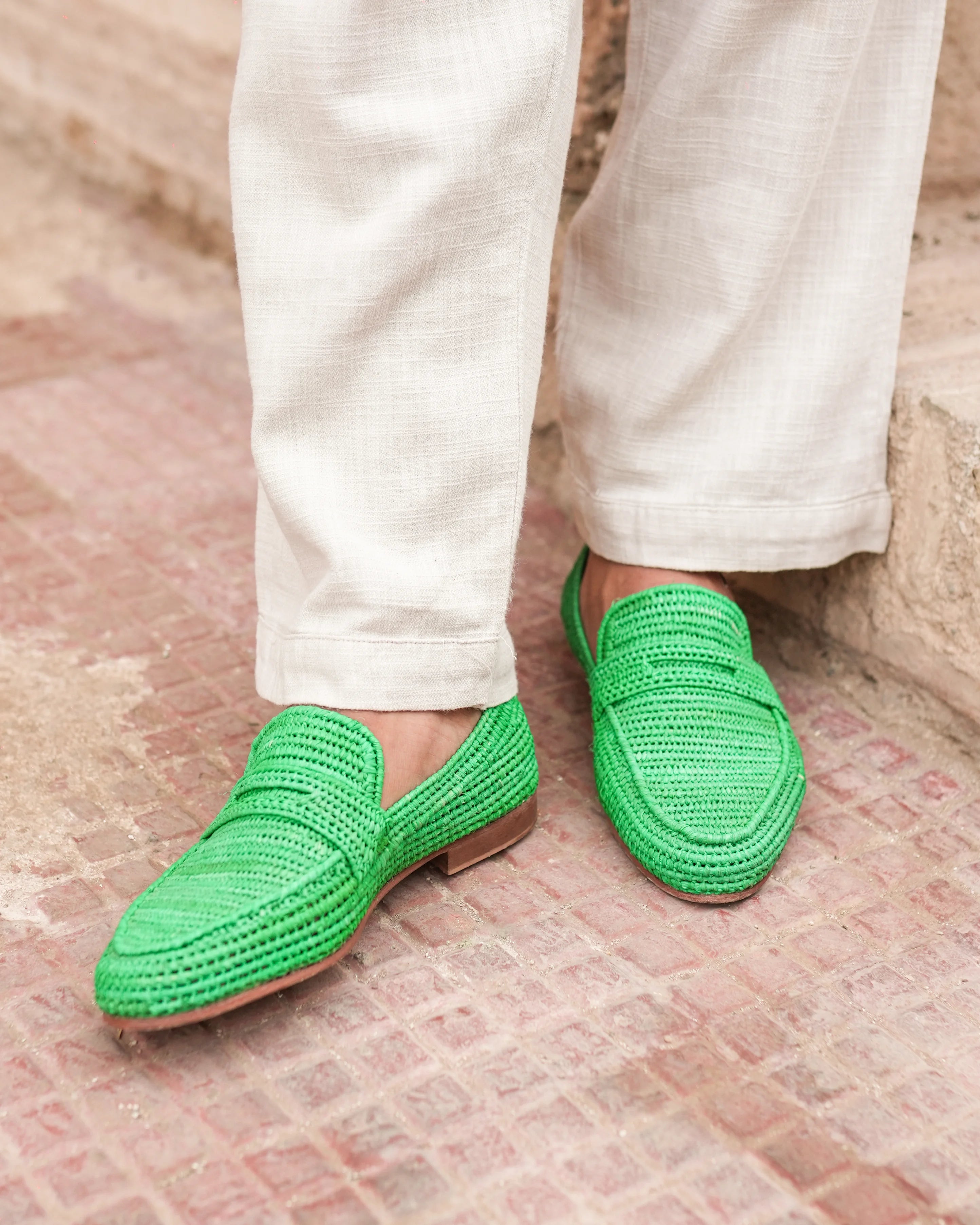 Green woven loafers worn with white pants on a textured floor.
