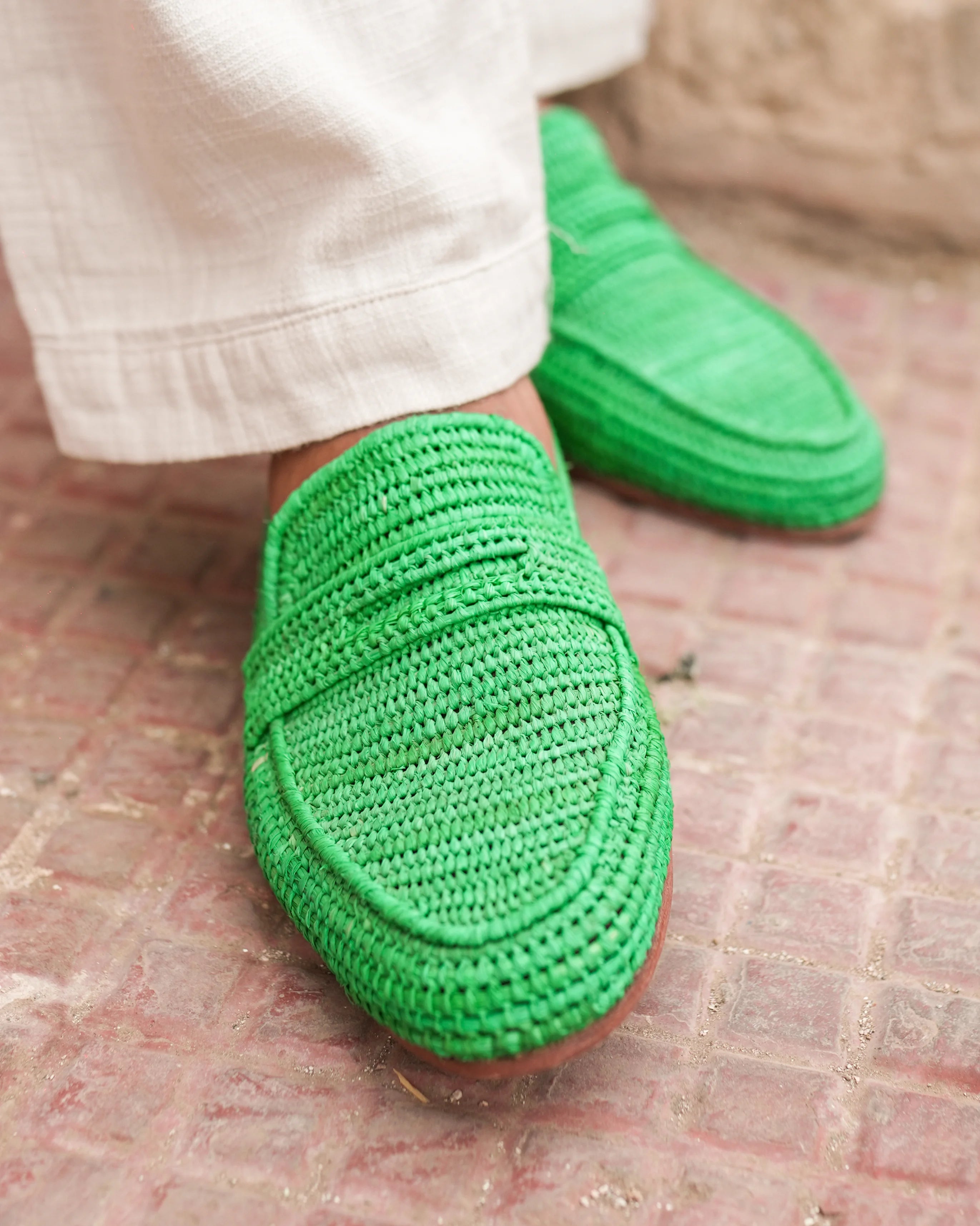 Green crochet loafers worn with white pants on a textured floor.