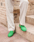 Green raphia shoes worn with white pants on stone steps.