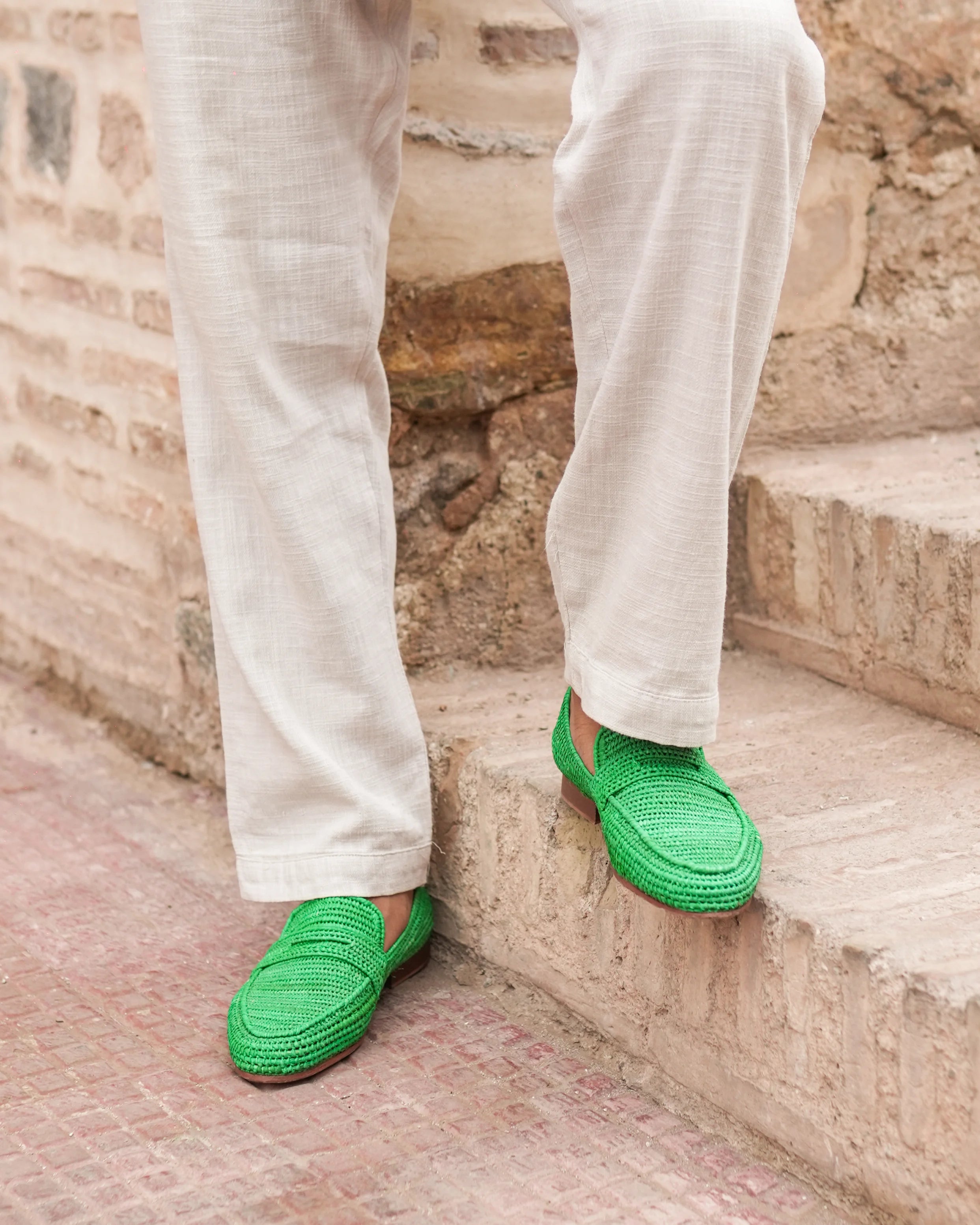 Green raphia shoes worn with white pants on stone steps.