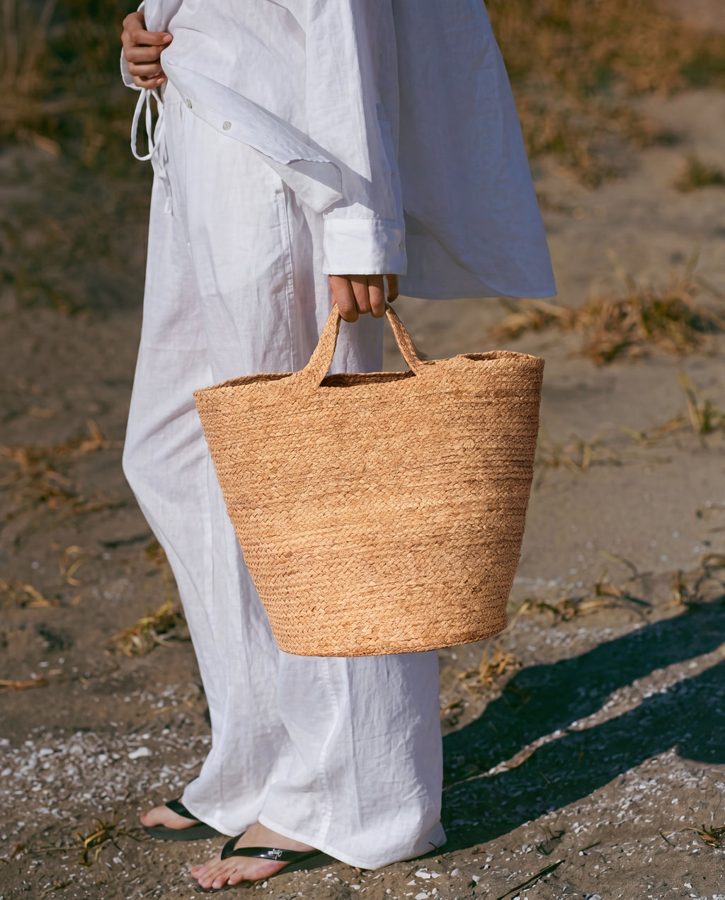 A person in a white linen outfit carrying a large natural woven straw tote bag on a sandy beach path.