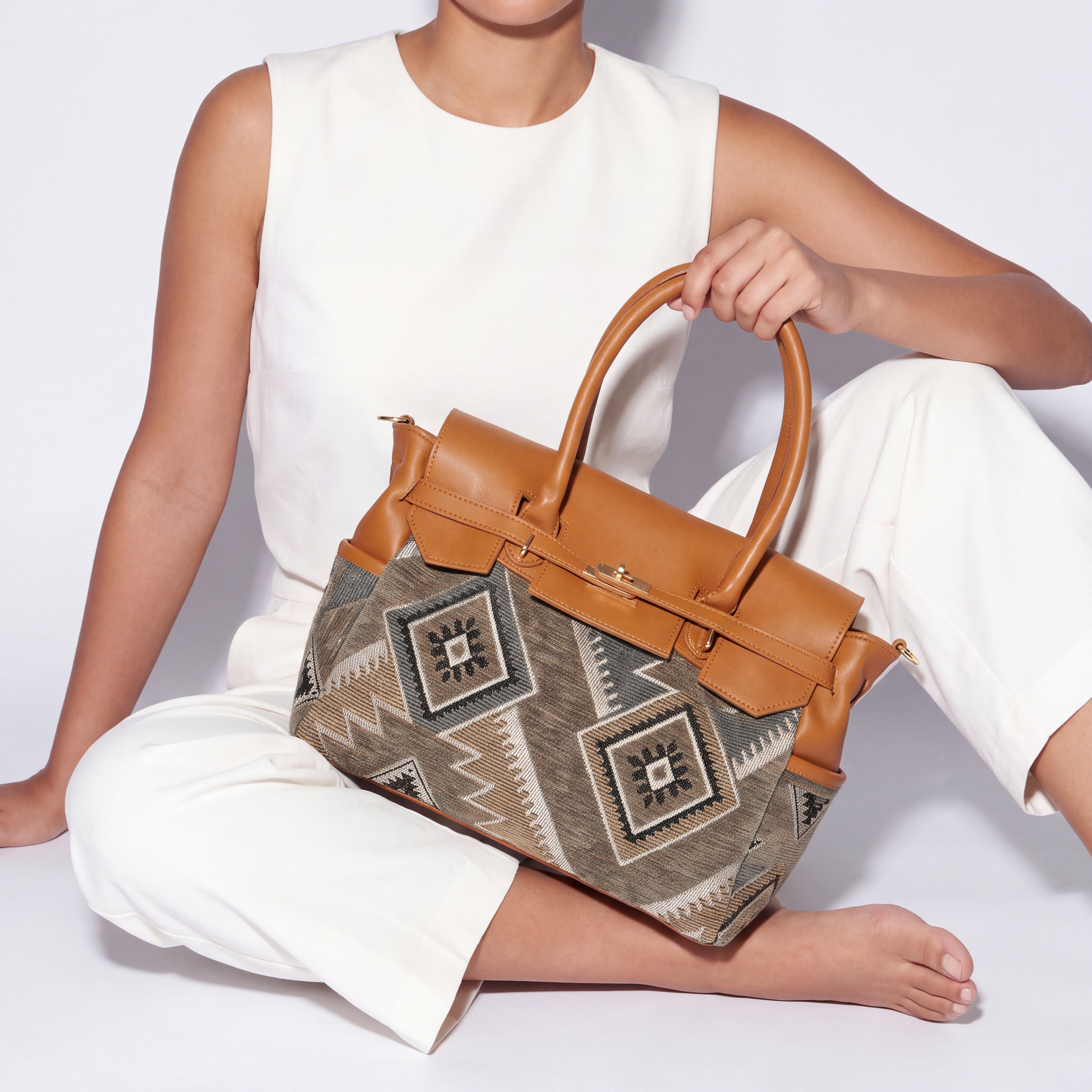 A woman in a white sleeveless top and white pants sits on a white floor holding a structured handbag. The bag has tan leather trim and handles with a geometric patterned front panel in beige, gray, and brown tones.