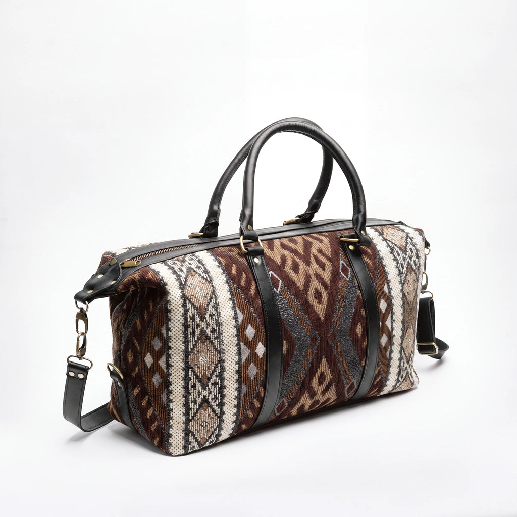 A duffle bag with a brown, beige, and gray geometric patterned fabric body, black leather handles and trim, and gold-tone hardware, photographed on a white studio background.