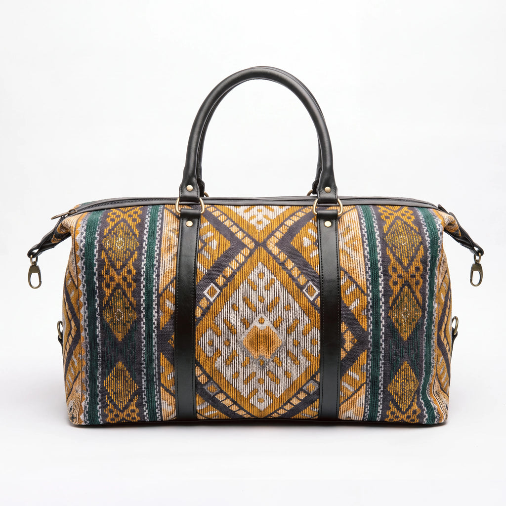 A structured duffel bag with a geometric ikat pattern in mustard yellow, brown, and dark teal, featuring black leather handles and vertical straps with gold-tone hardware, photographed against a plain white background.