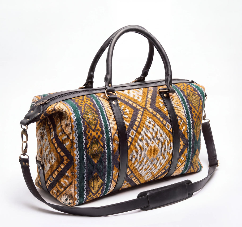 A duffel bag with a colorful geometric patterned fabric body and black leather handles, trim, and adjustable shoulder strap, photographed on a clean white background.