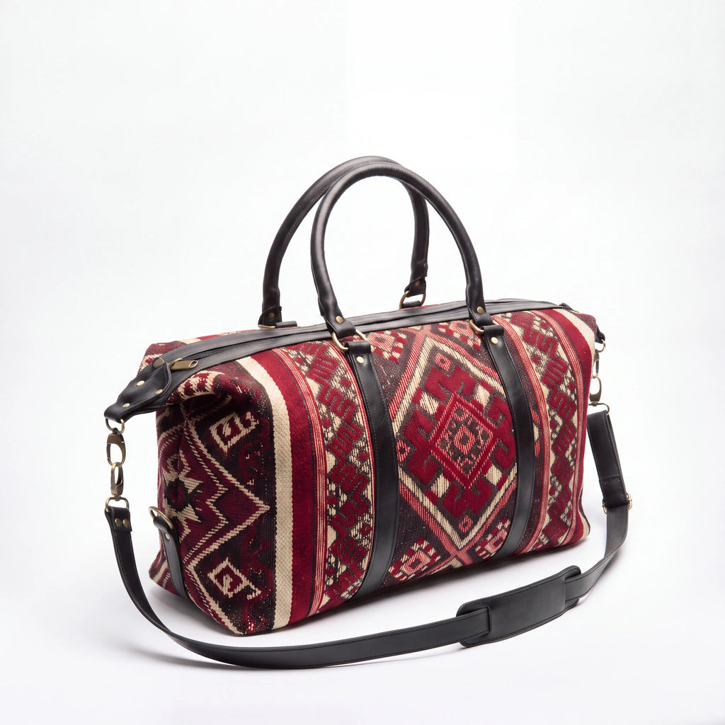 A duffel bag with a red and beige geometric kilim pattern, accented by black leather handles and a matching black shoulder strap, set against a plain white background.