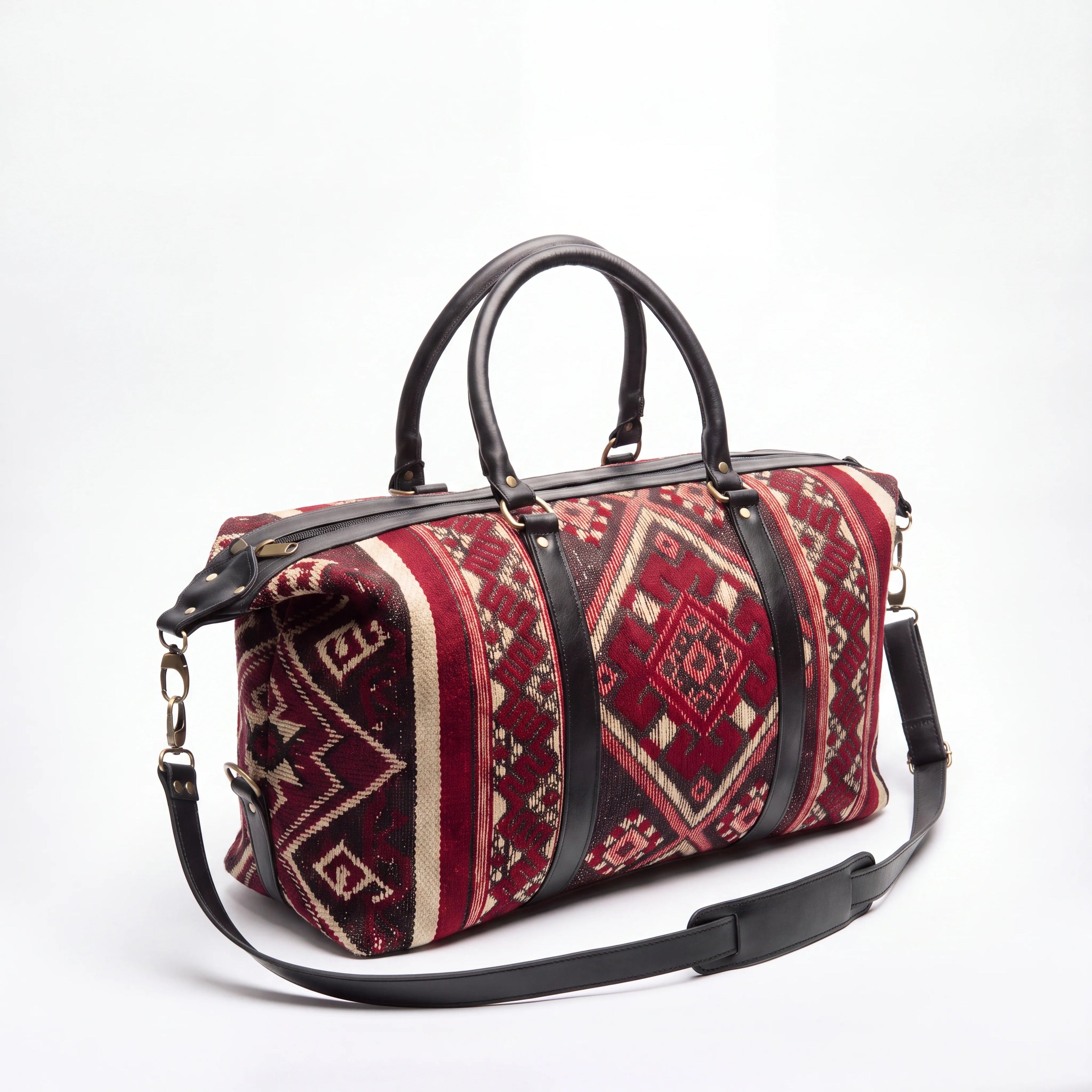 A duffel bag with a red and beige geometric kilim pattern, accented by black leather handles and a matching black shoulder strap, set against a plain white background.