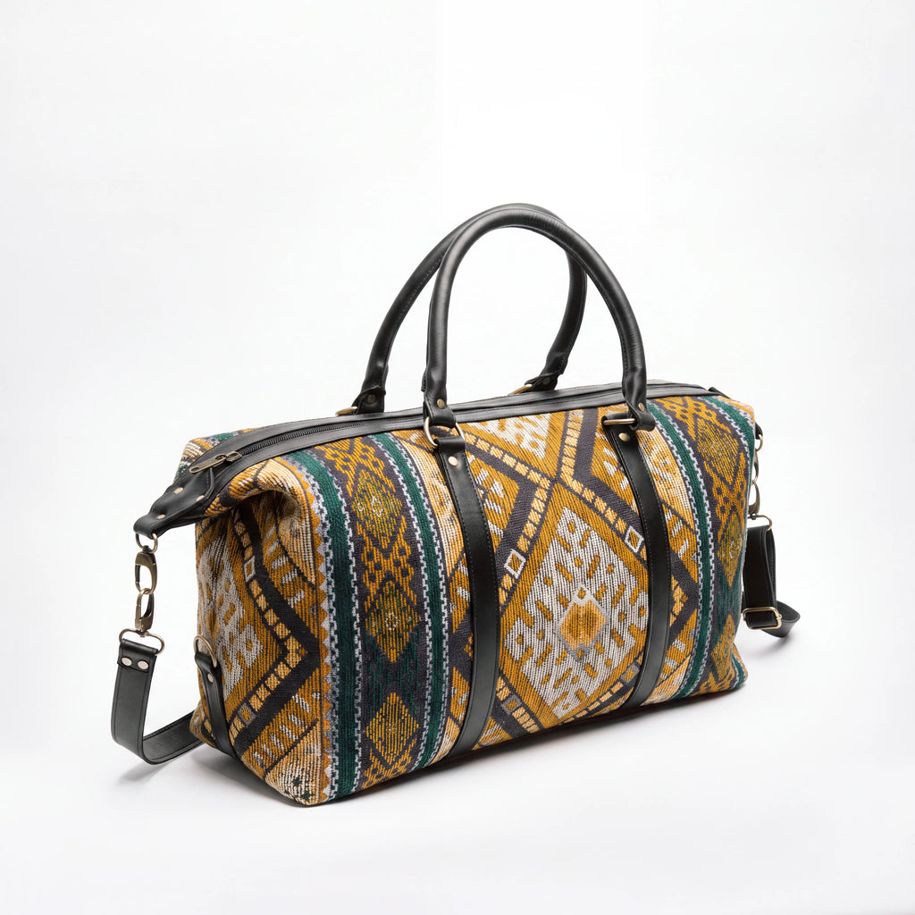 A duffle bag with a geometric patterned fabric in mustard yellow, beige, and green, accented with black leather handles and straps, shown on a white background.