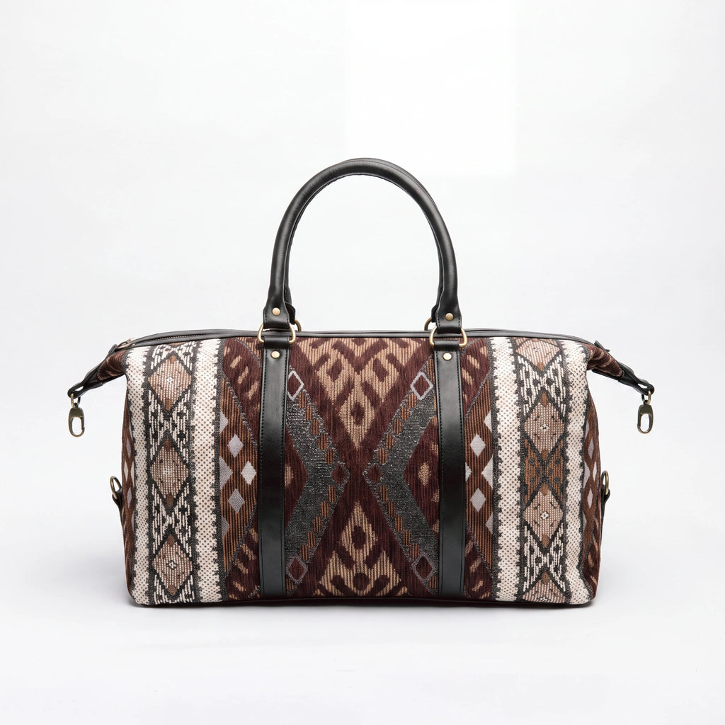 A duffle bag with a brown, beige, and black geometric patterned fabric body, featuring two black leather top handles and black vertical straps with gold-tone hardware, set against a plain white background.