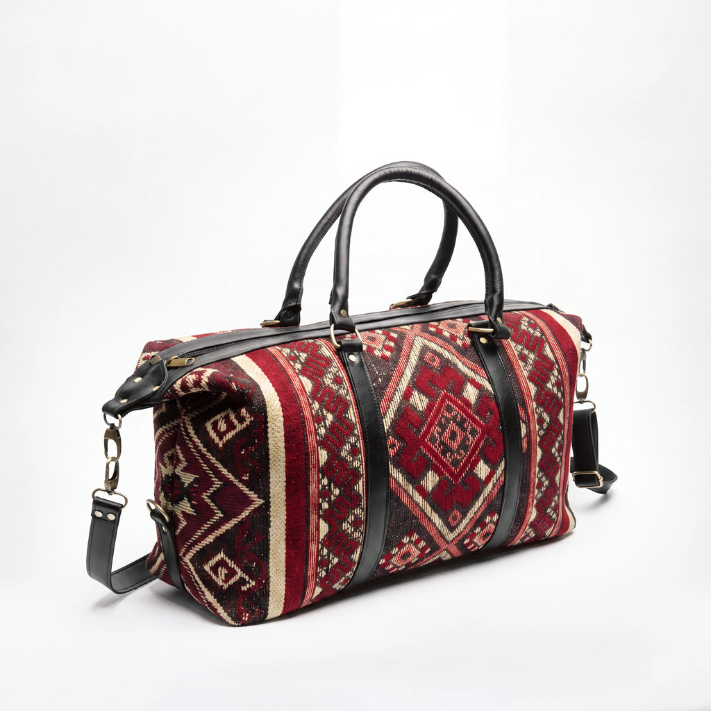 A duffel bag with a red, beige, and black geometric patterned fabric body, featuring black leather handles and trim, photographed on a white studio background.