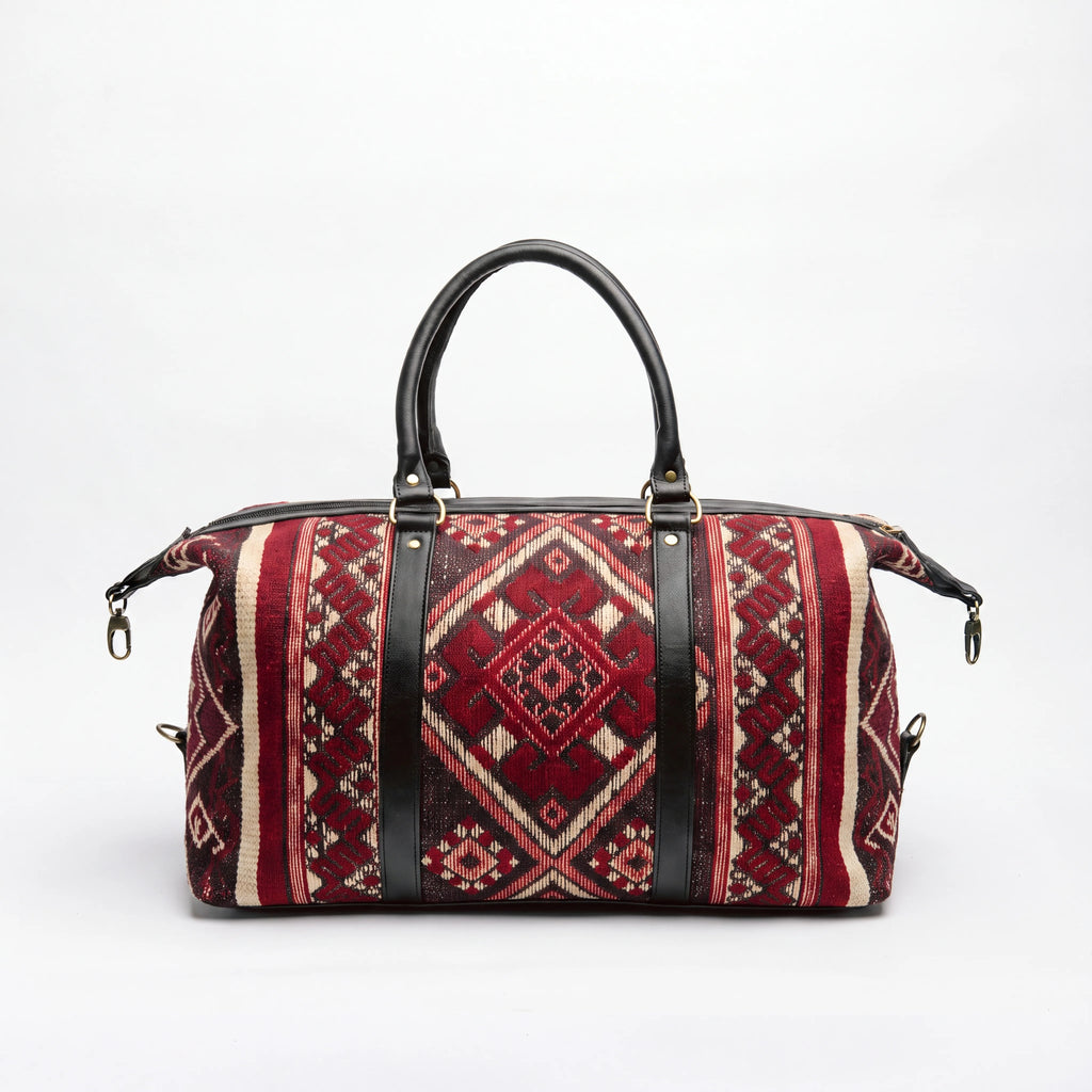 A duffel bag with a red and beige geometric kilim pattern and black leather handles and straps, shown on a white background.