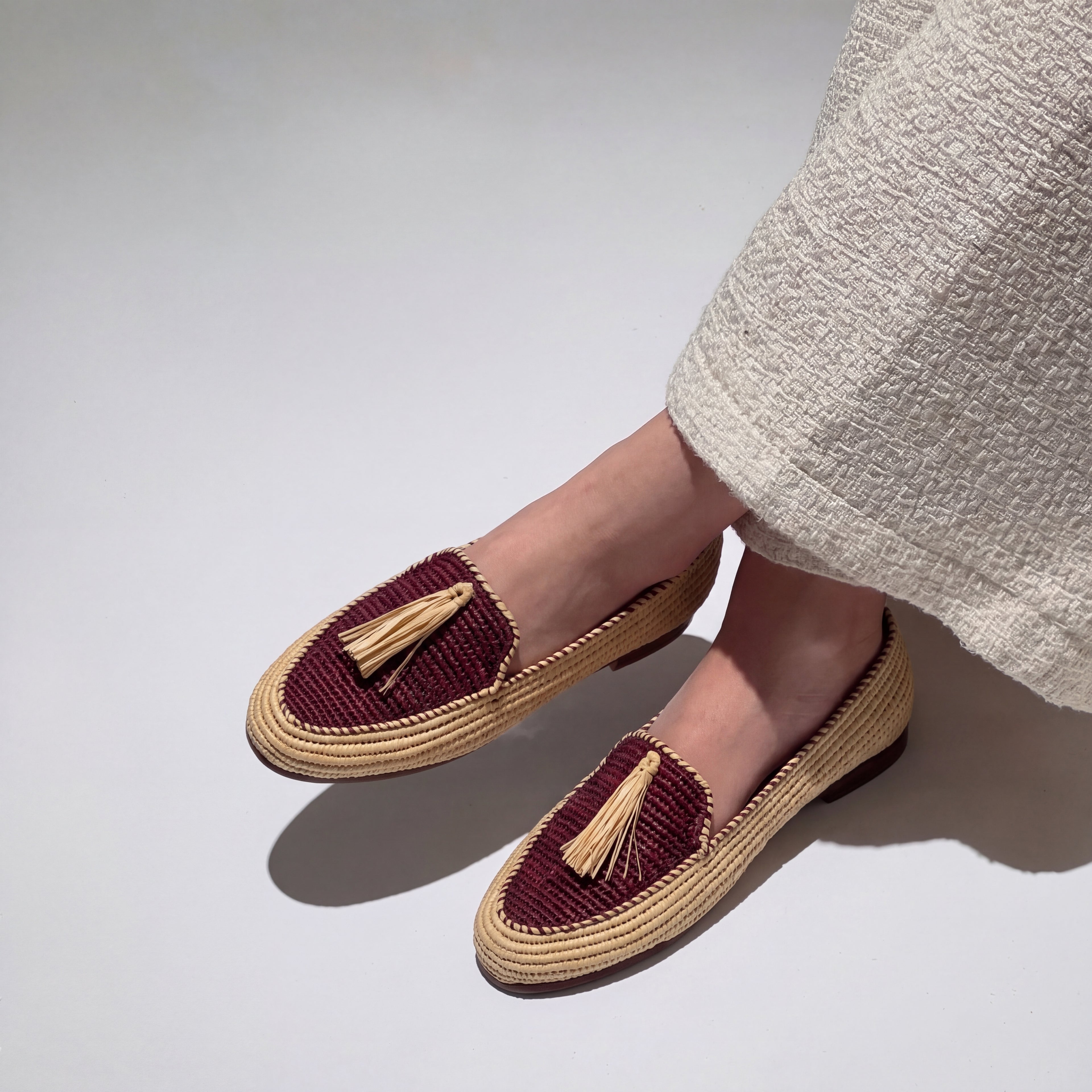 A person wearing woven loafer-style flats with deep burgundy uppers and beige tassel details, paired with a light beige textured skirt, shown against a plain light gray studio background.