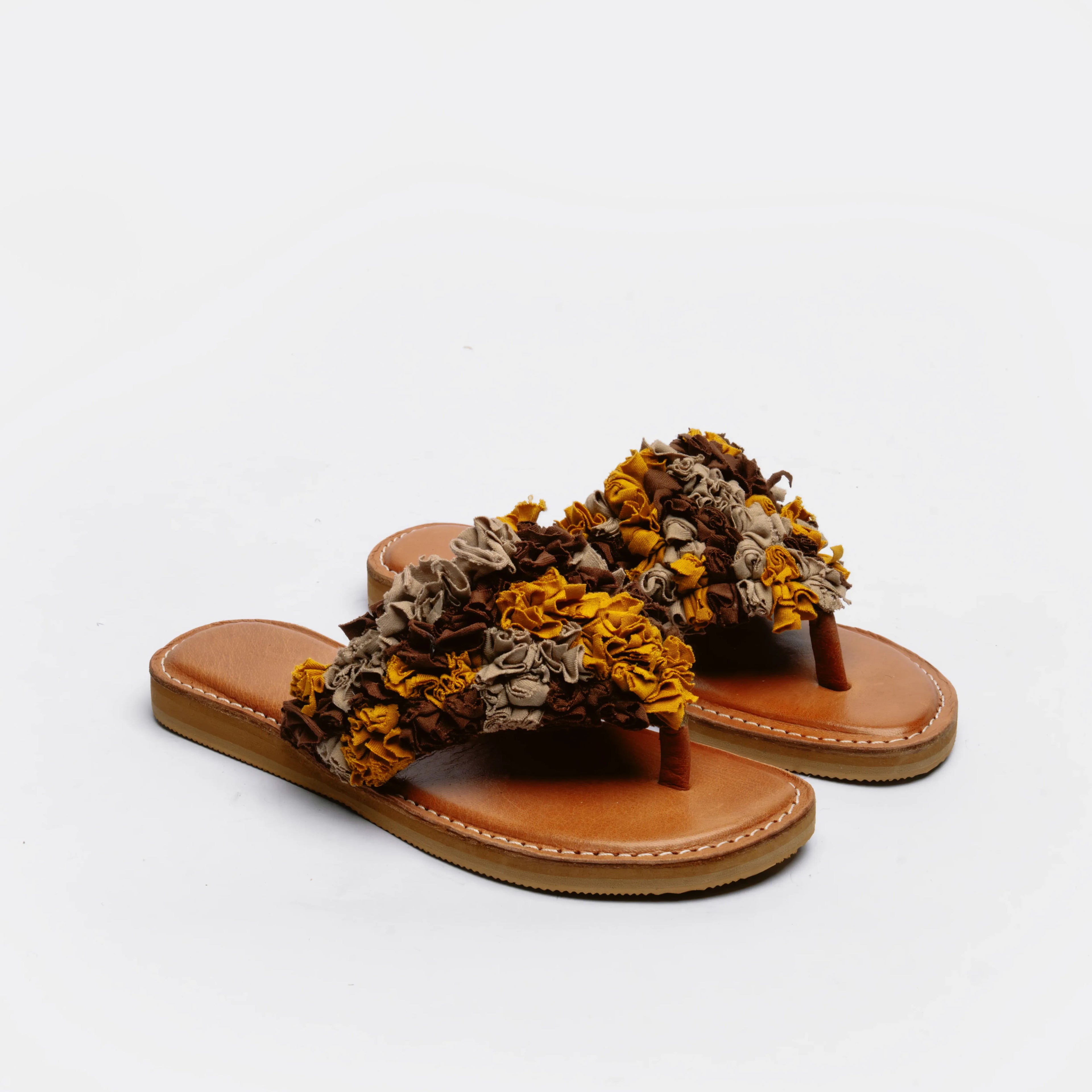 A pair of thong sandals with tan leather footbeds and straps adorned with textured, ruffled dried flowers in mustard yellow, brown, and beige, set against a plain white background.