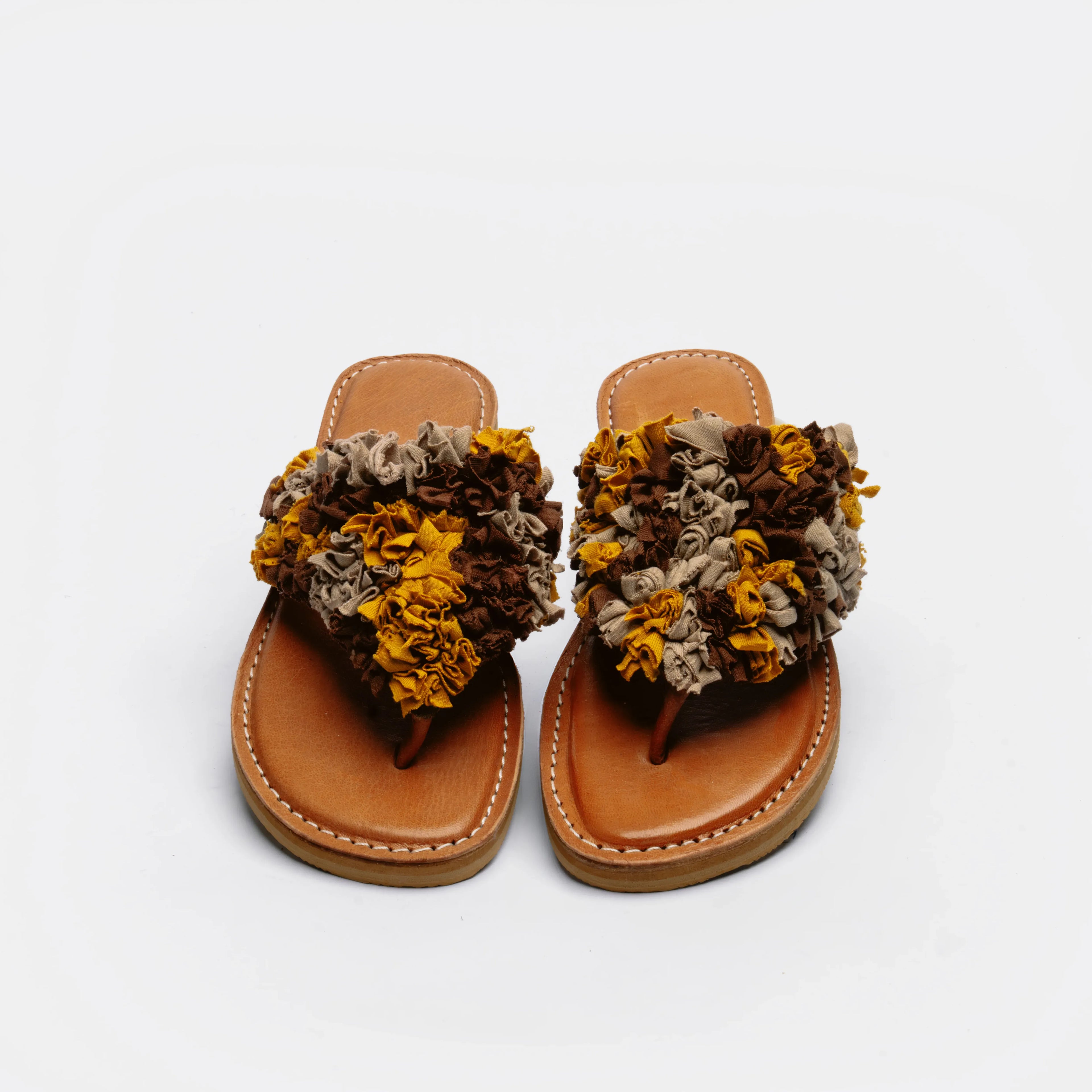 A pair of tan leather thong sandals with large, decorative ruffled straps in brown, beige, and mustard yellow, photographed on a clean white background.