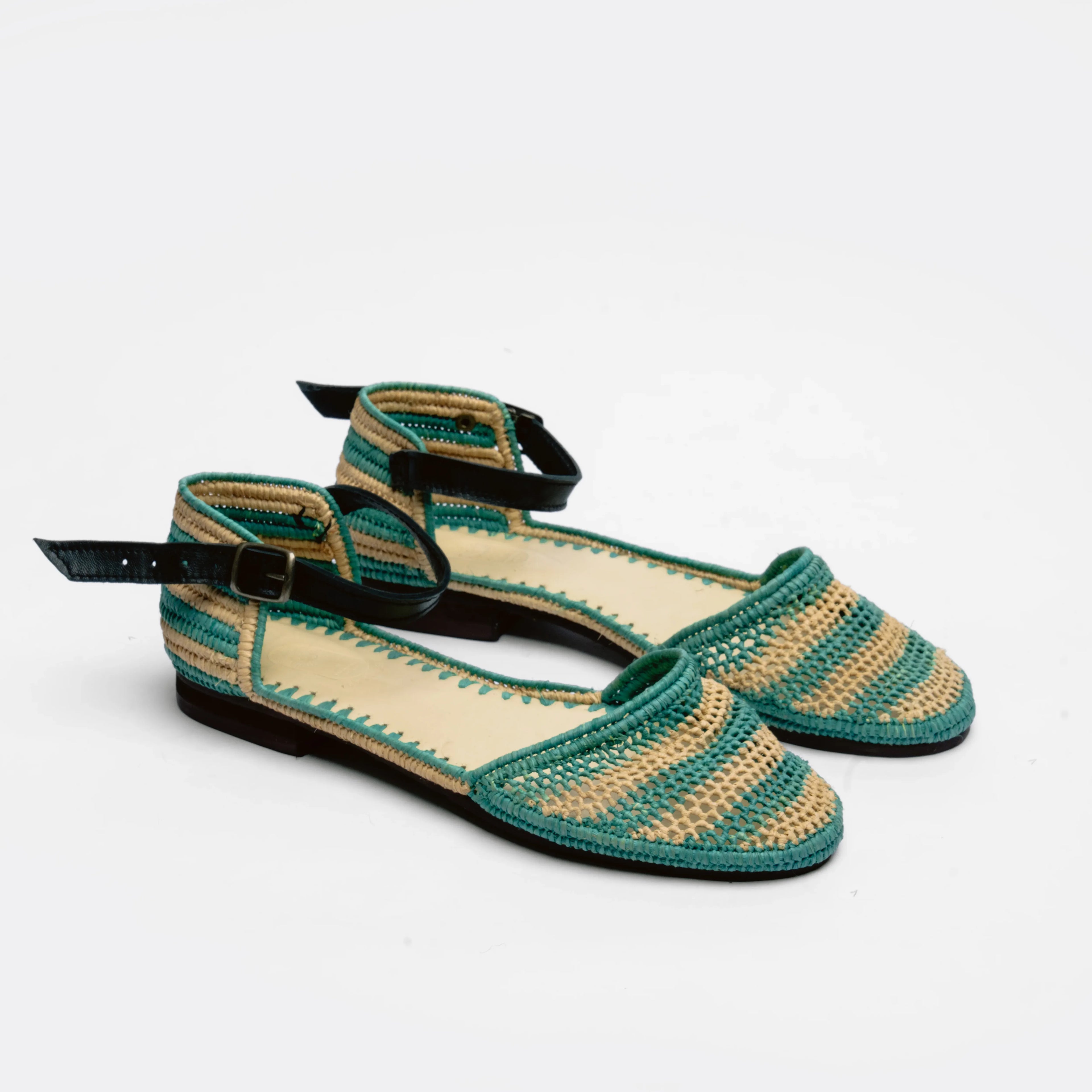 A pair of woven flat sandals with green and beige horizontal stripes, black ankle straps with gold-tone buckles, and a low black heel, shown on a white background.