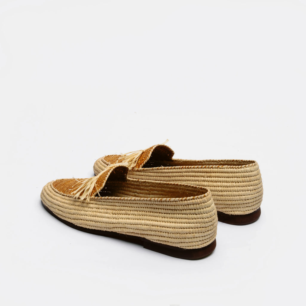 A pair of woven raffia slip-on loafers with tassel details, shown from a side-rear angle on a clean white background.