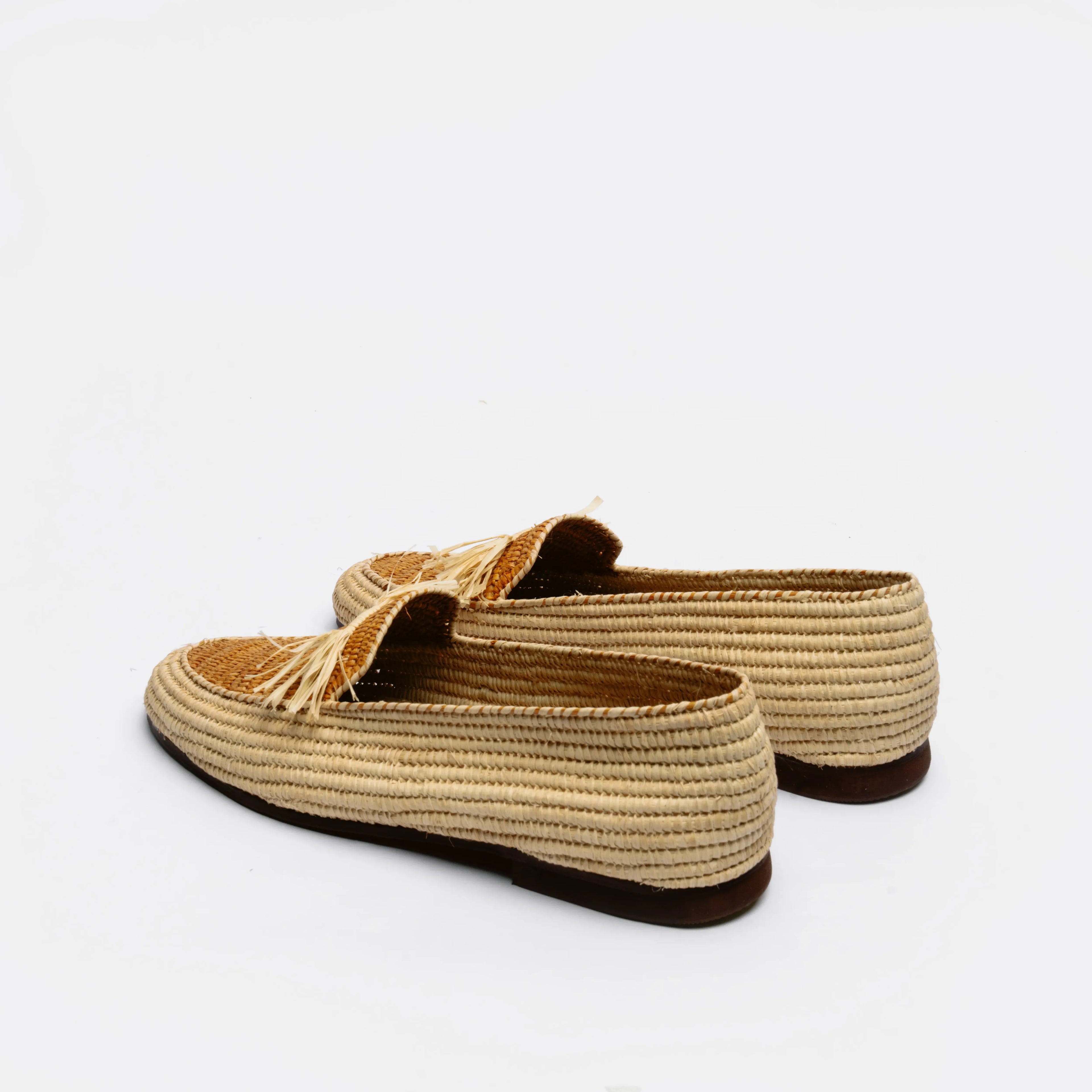 A pair of woven raffia slip-on loafers with tassel details, shown from a side-rear angle on a clean white background.